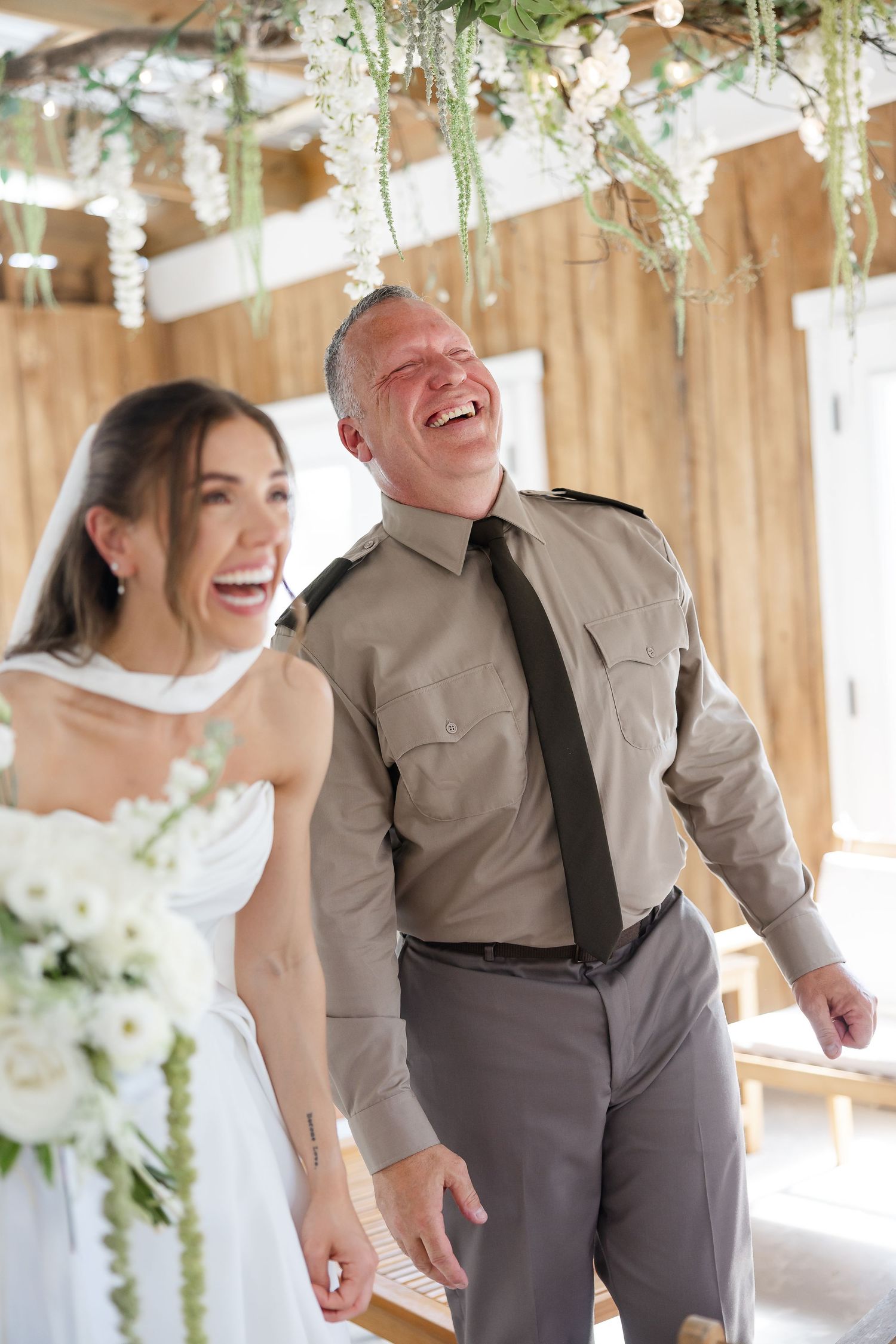 Bride in white dress and man in military uniform laughing joyfully at a wedding ceremony with floral decorations.