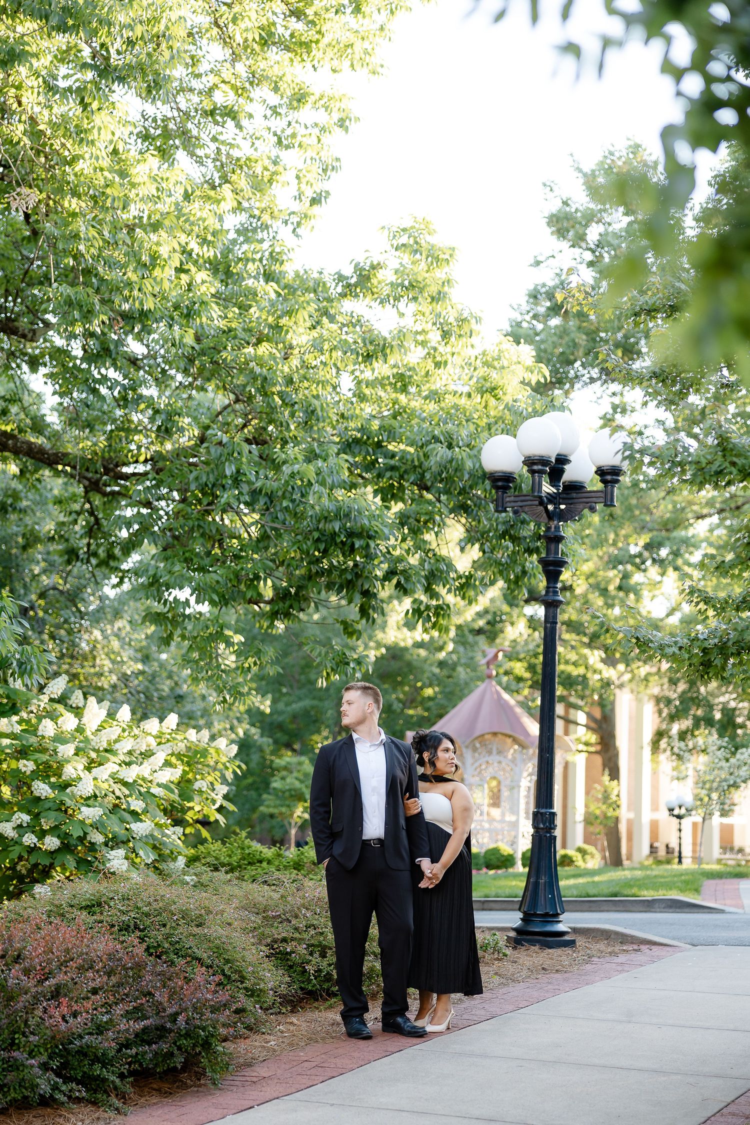Couple in formal attire standing near a lamp post surrounded by lush green trees on a brick pathway.