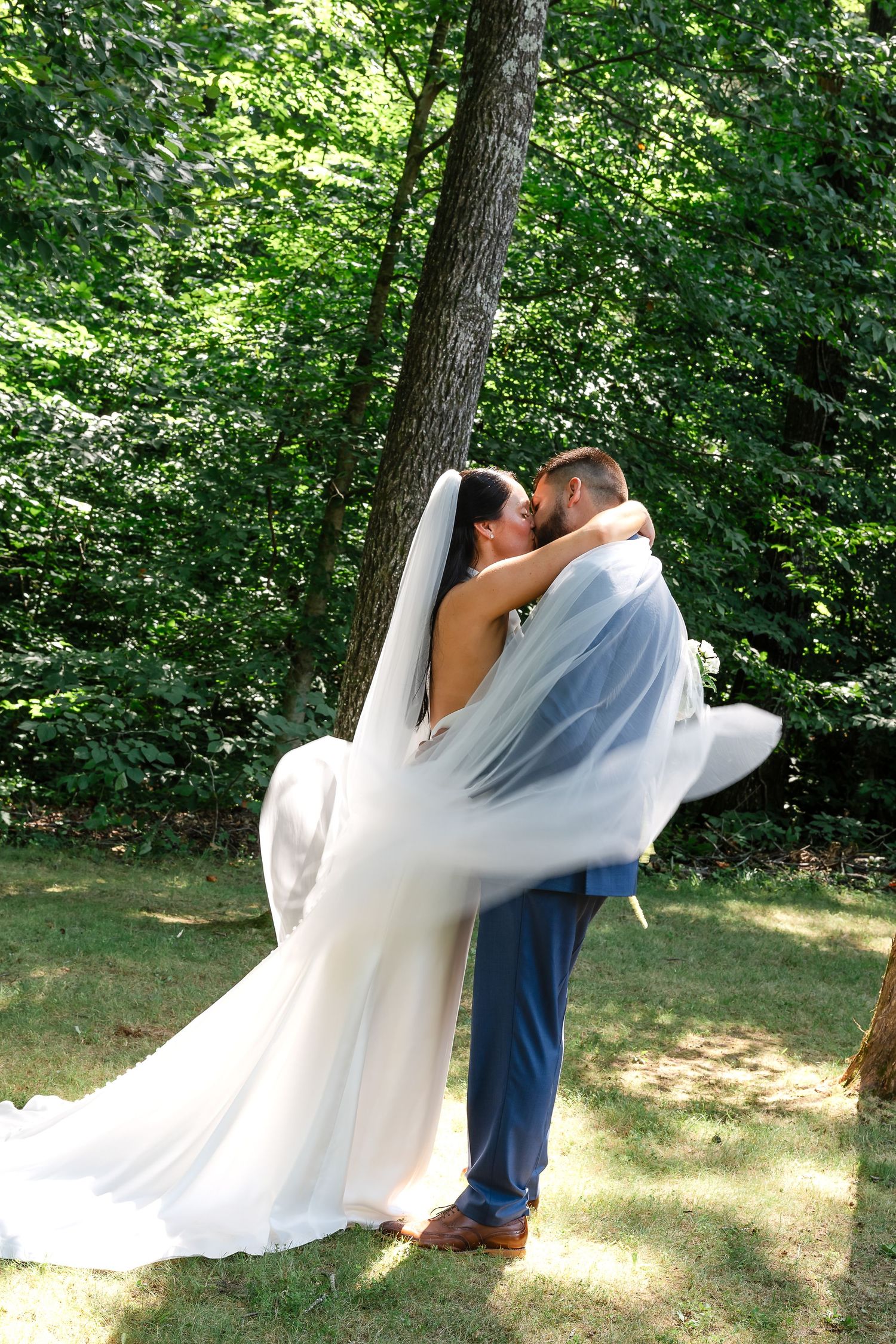 Groom lifts bride in white gown and veil, sharing a romantic kiss outdoors surrounded by lush green trees.