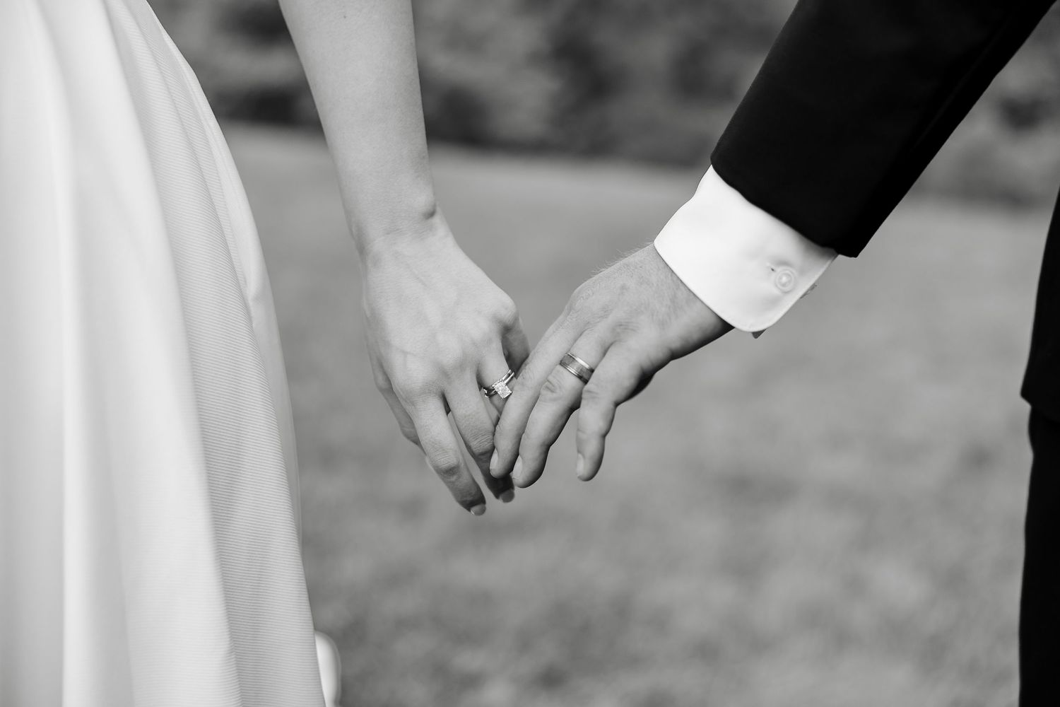 Black and white photo of a bride and groom holding hands, showing wedding rings, in an outdoor setting.