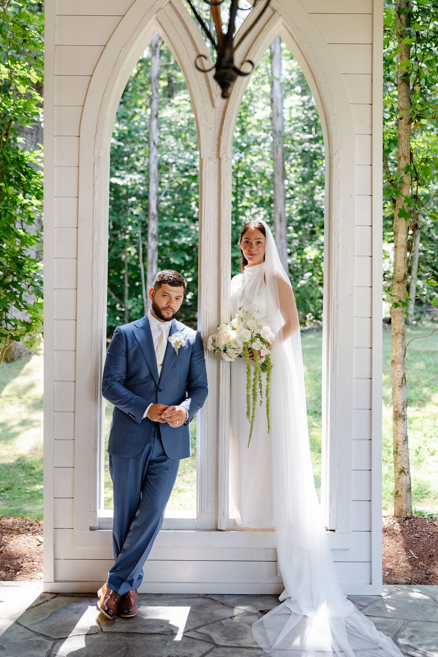 Bride and groom pose in a white arched chapel gazebo, she in a veil with bouquet, he in a blue suit. at firefly lane in Dickson, tn