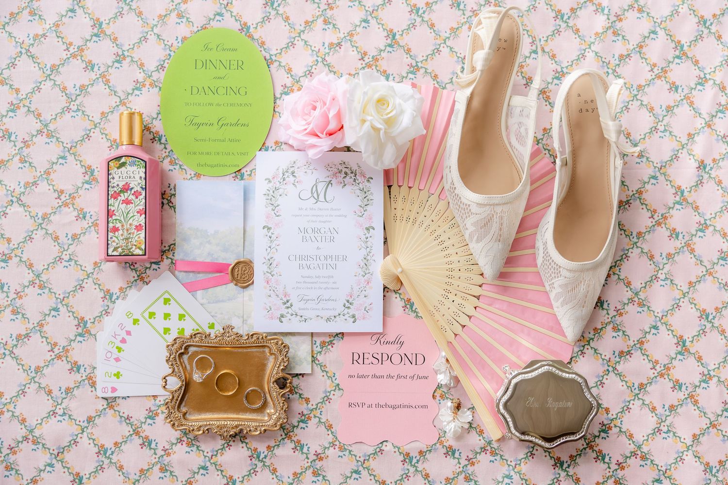Elegant wedding flat lay featuring floral invitations, pink fan, lace heels, gold rings, perfume, and pink peony on floral backdrop.
