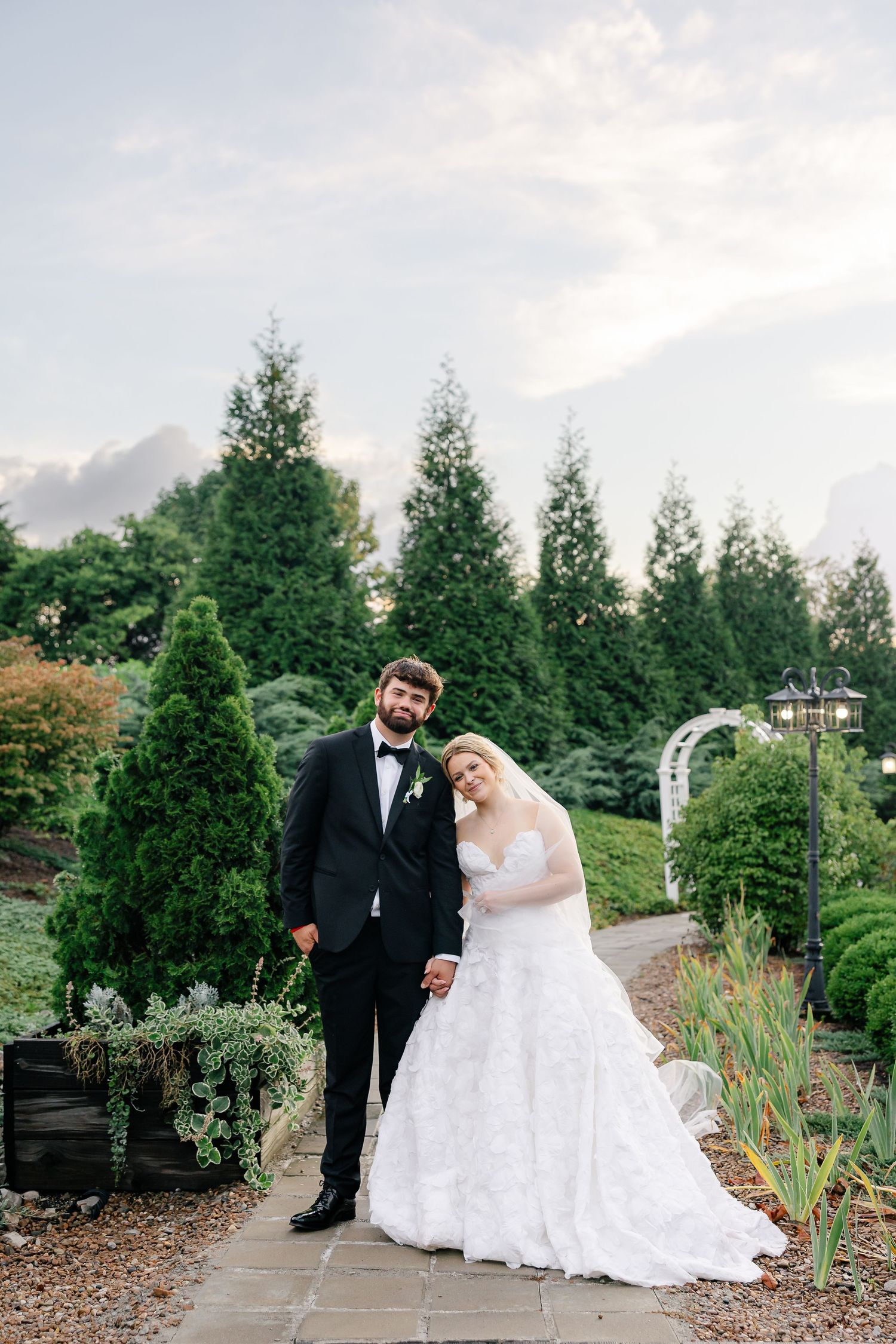 Bride and groom pose on garden path surrounded by evergreen trees, she in lace gown, he in black tuxedo.