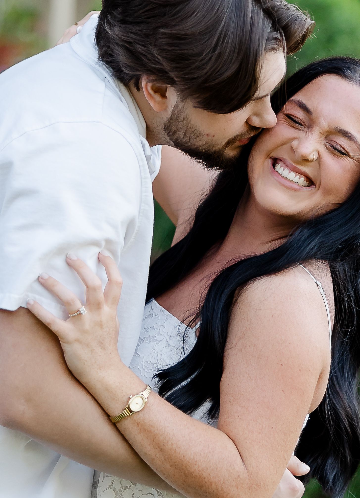 Happy couple embracing outdoors, man kissing smiling dark-haired woman wearing a gold watch.