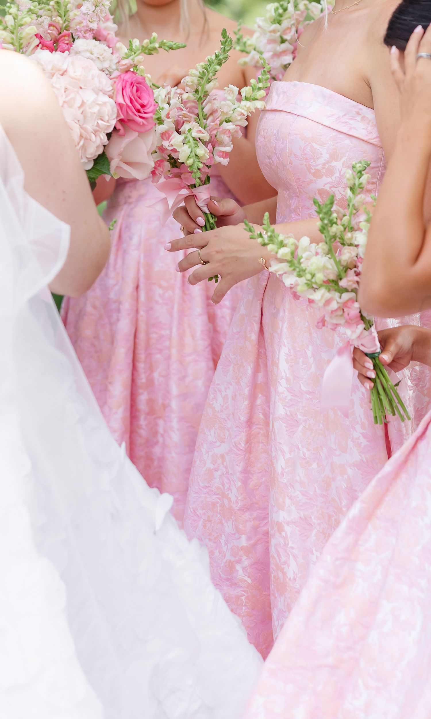 Bridesmaids in pink floral strapless gowns holding bouquets of pink roses and snapdragons at a wedding.