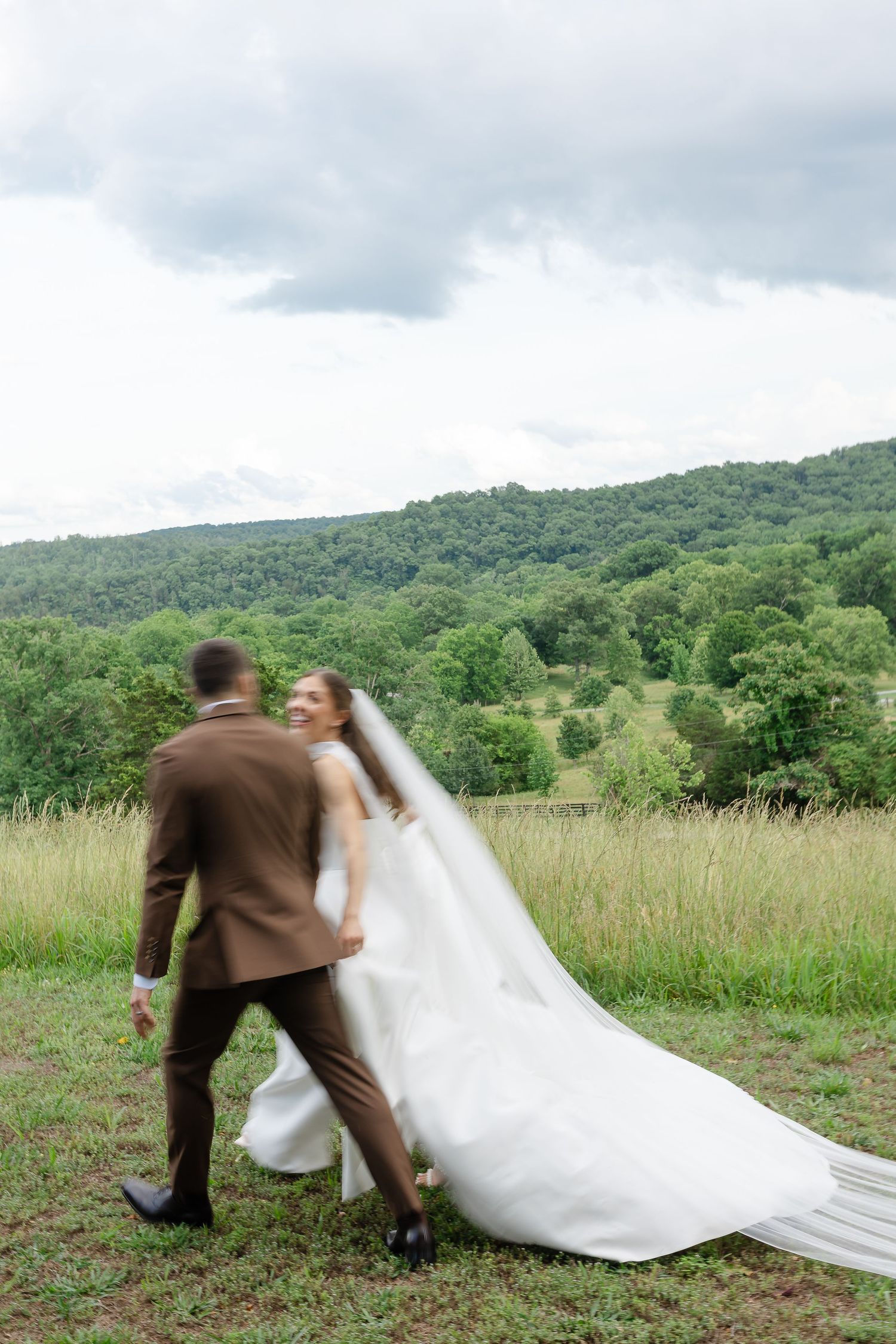 Bride and groom walk hand in hand through a lush green meadow with forested hills backdrop on their wedding day.