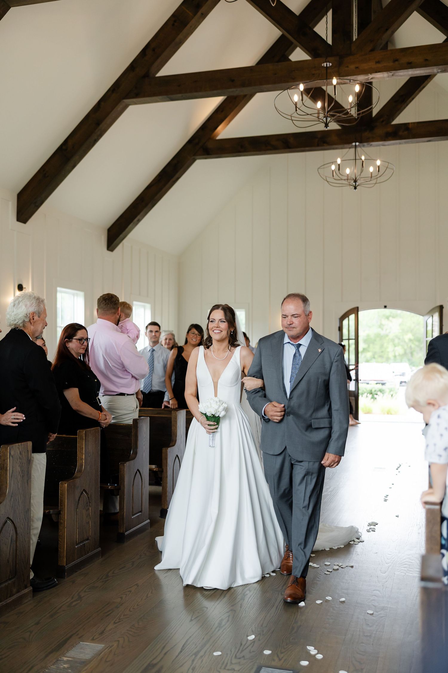 Bride walks down the aisle with her father in a white chapel with wooden beam ceilings during wedding ceremony. At The Venue at Birchwood
