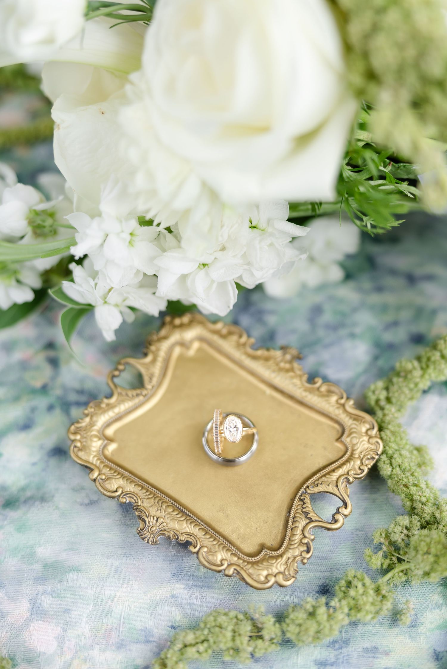 Diamond engagement ring displayed on gold ornate tray beside white roses and greenery for wedding flat lay.
