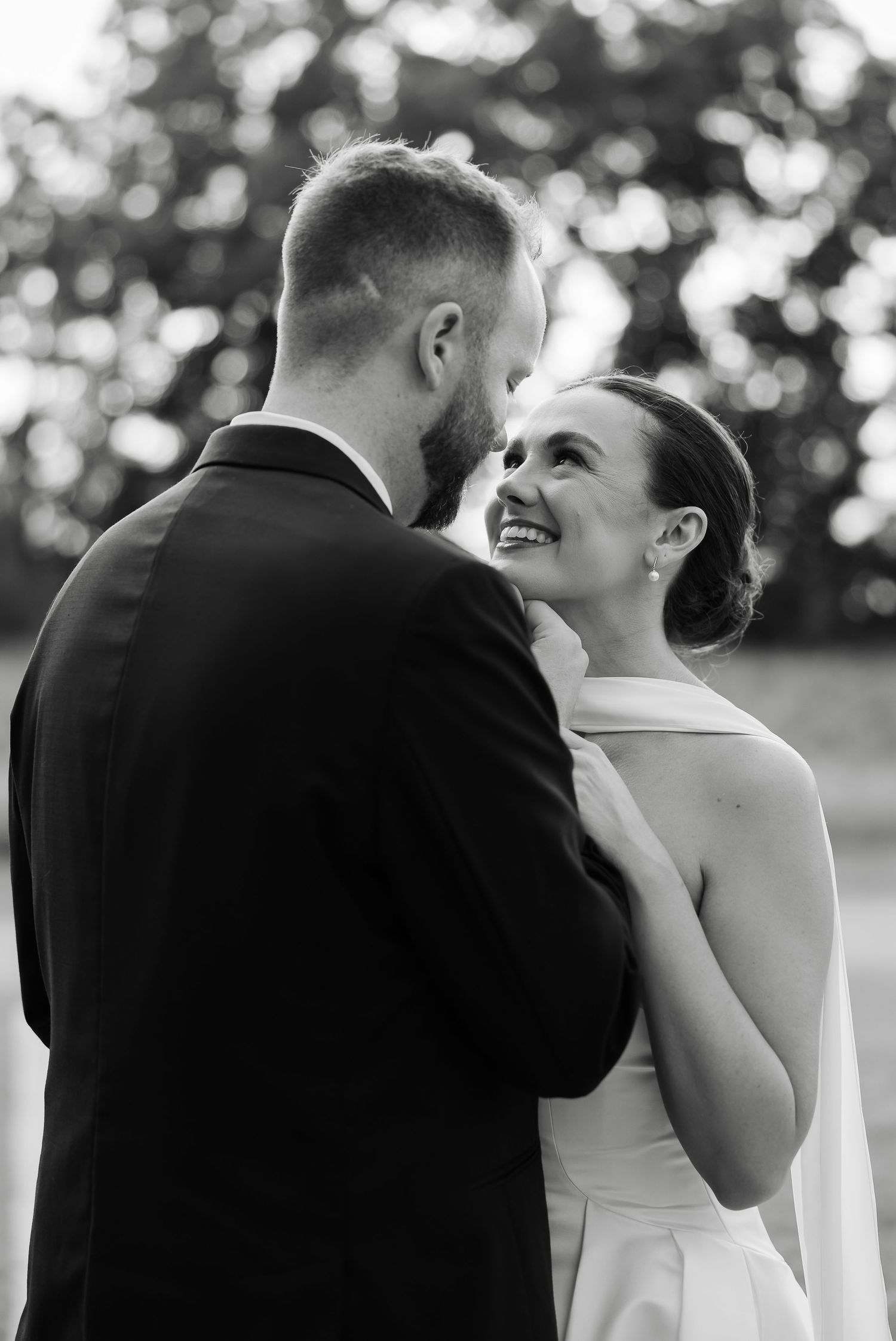 Black and white photo of a smiling bride in a white dress gazing up at her groom in a black suit outdoors. at the venue at birchwood