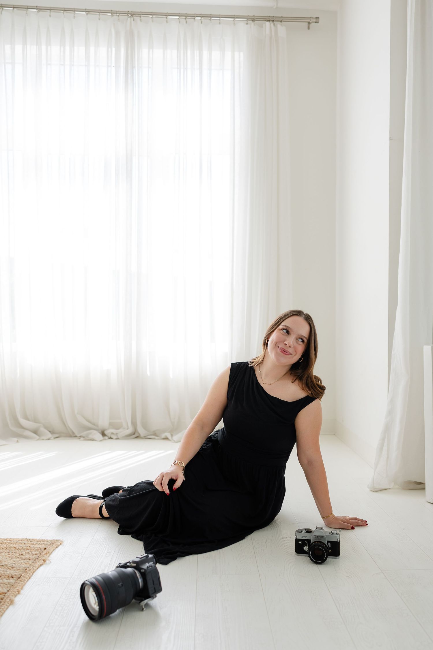 Woman in black dress sitting on white floor with DSLR and film cameras in bright studio setting.