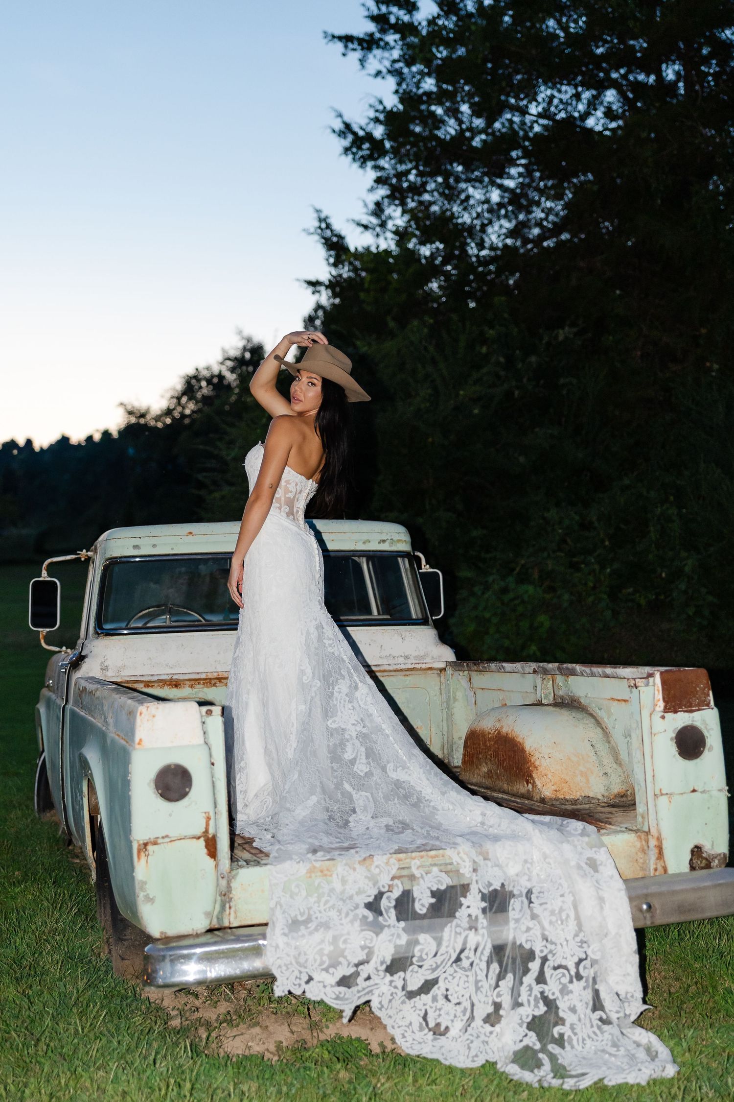Bride in wedding gown and cowboy hat poses on vintage rusty pickup truck at dusk with trees in background At Drakewood Farms in Nashville