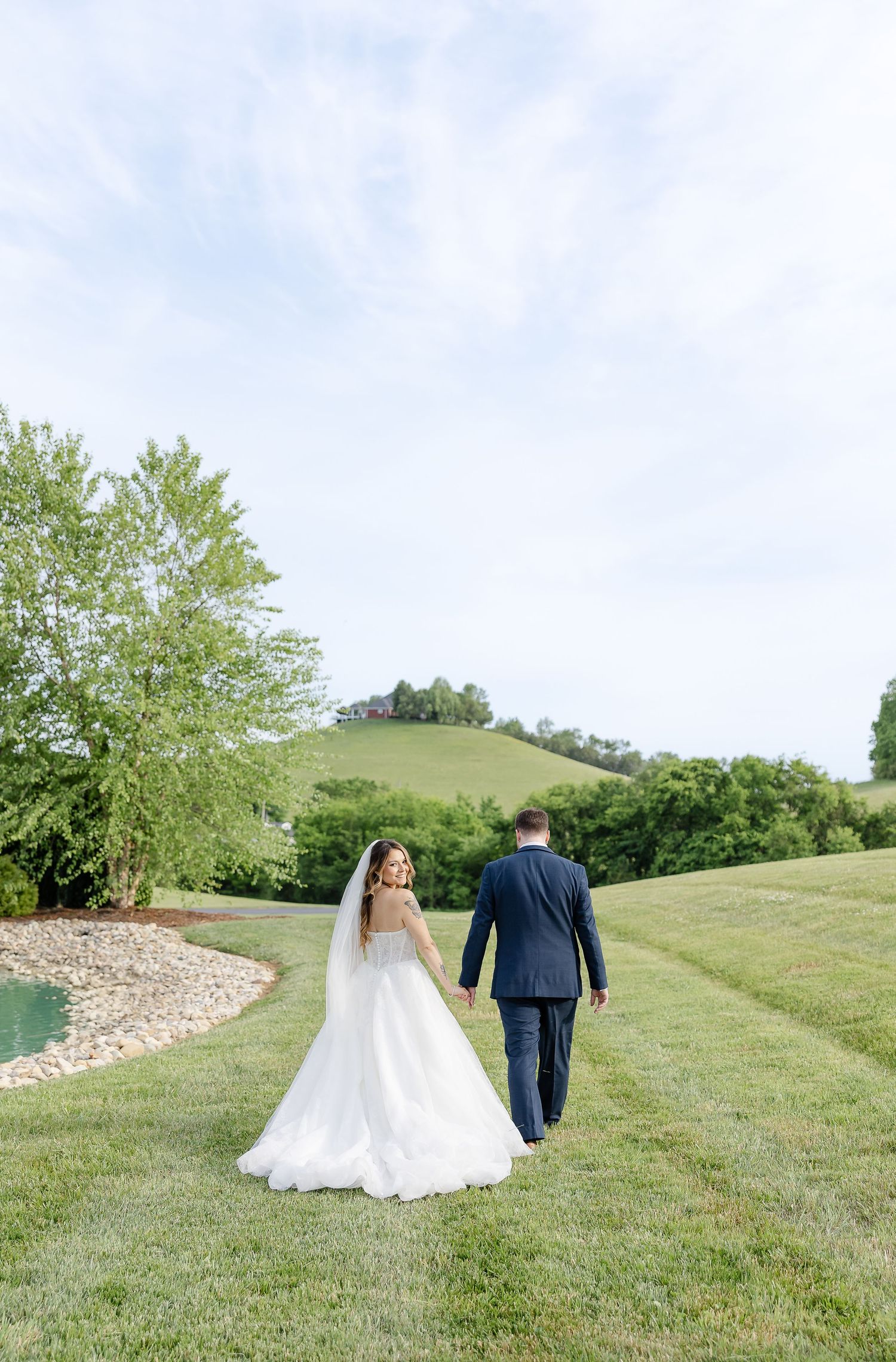 Bride and groom walk hand in hand across lush green hills at an outdoor wedding venue on a sunny day.