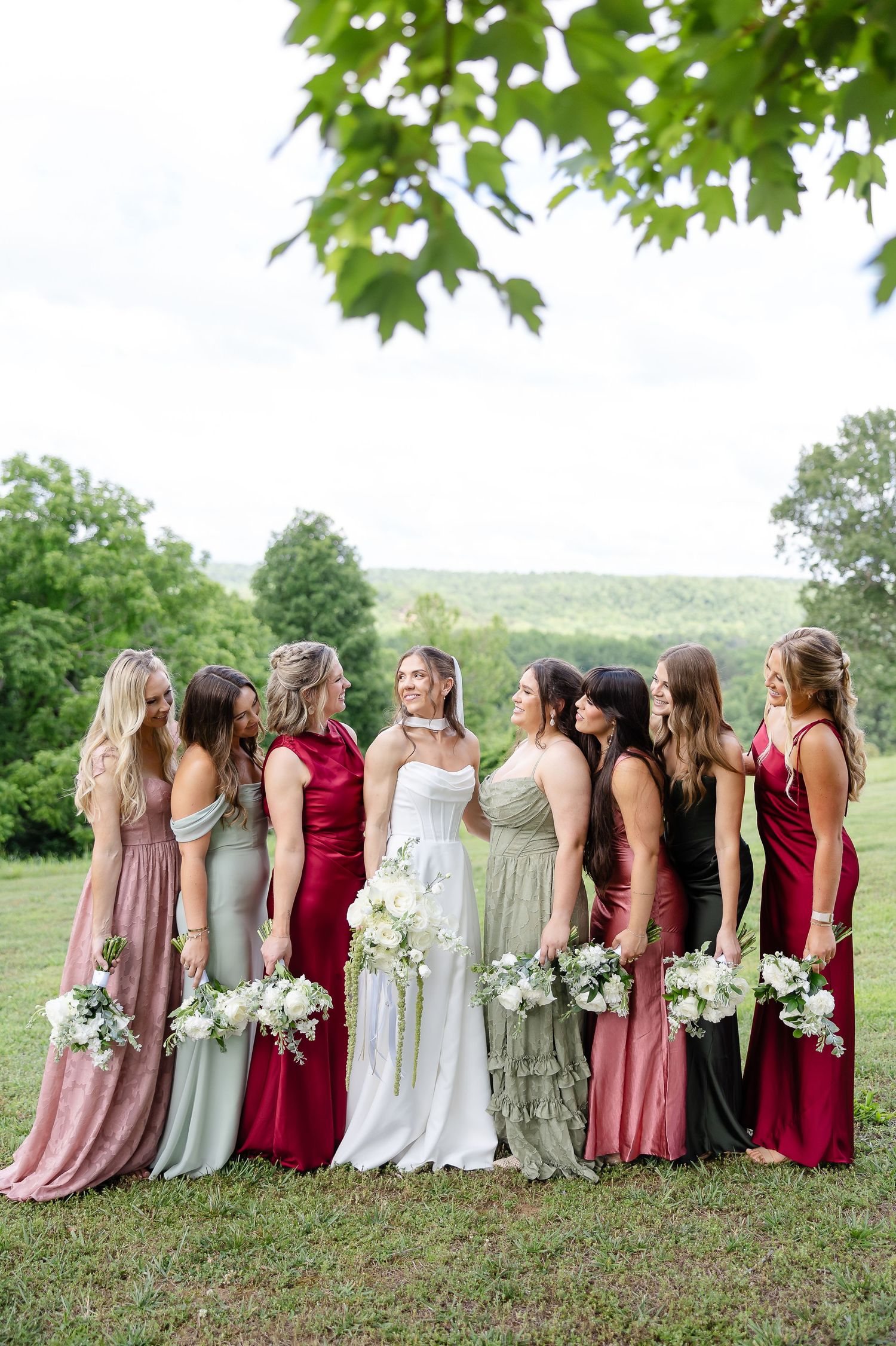 Bride poses with bridesmaids in mismatched pink and green gowns holding white bouquets outdoors.