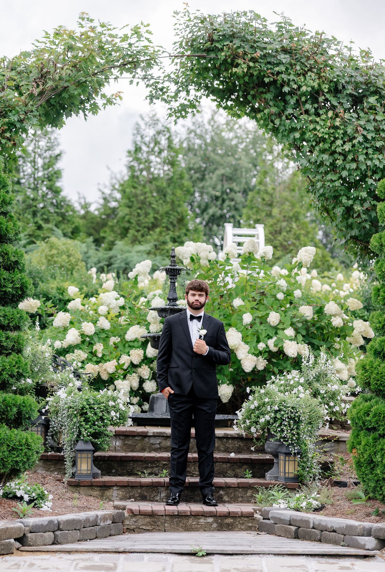 Groom in black tuxedo stands before white hydrangeas and garden arch at outdoor wedding ceremony.