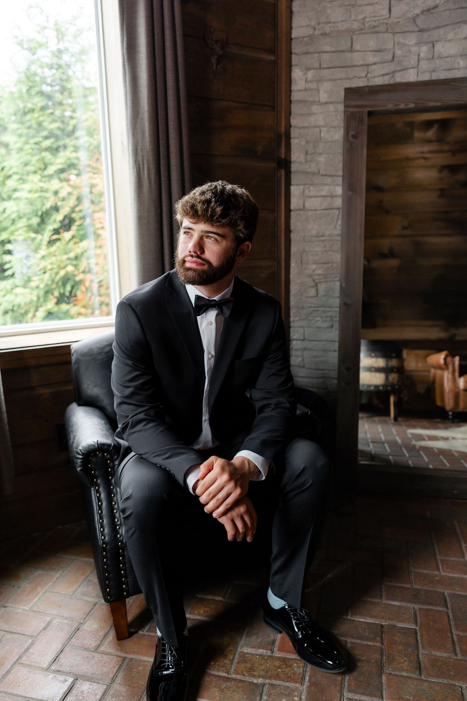A well-dressed groom in a black tuxedo and bow tie sits in a leather chair by a window near a stone fireplace.