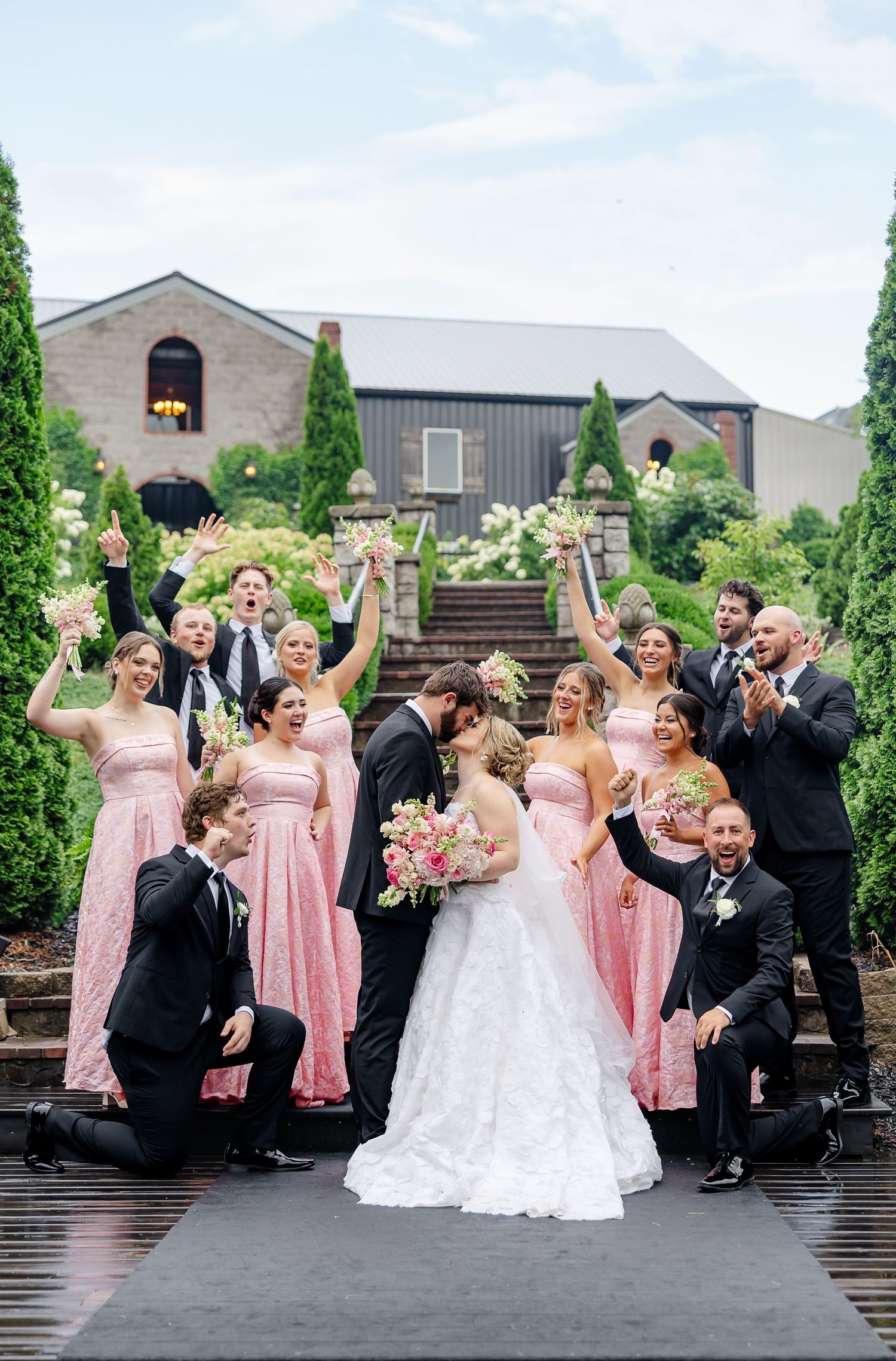 Wedding party celebrating outdoors, bridesmaids in pink gowns, groomsmen in black tuxedos, bride and groom kissing.