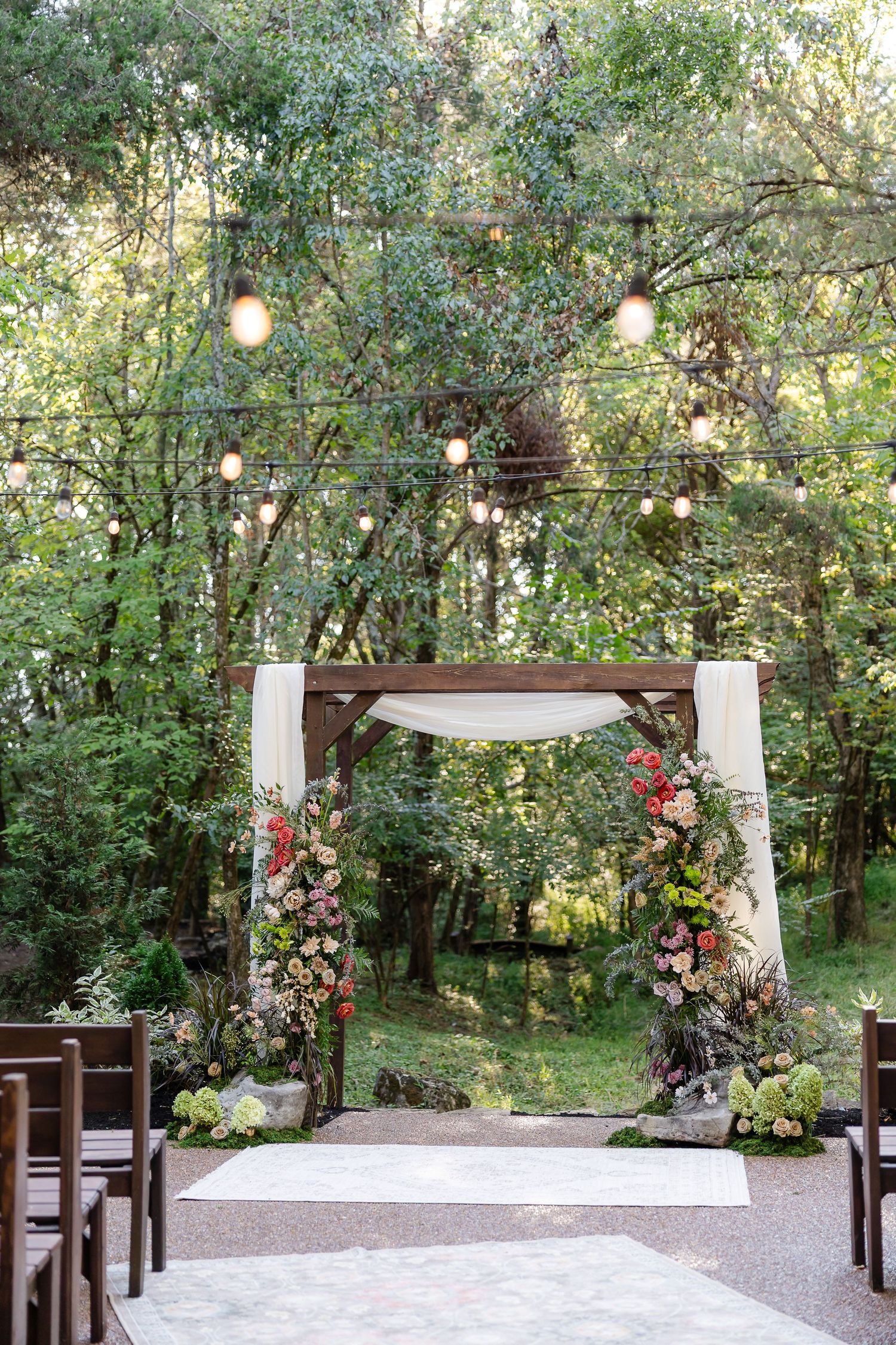 Outdoor wedding ceremony arch adorned with colorful flowers and white draping, surrounded by lush trees and string lights.At Drakewood Farms