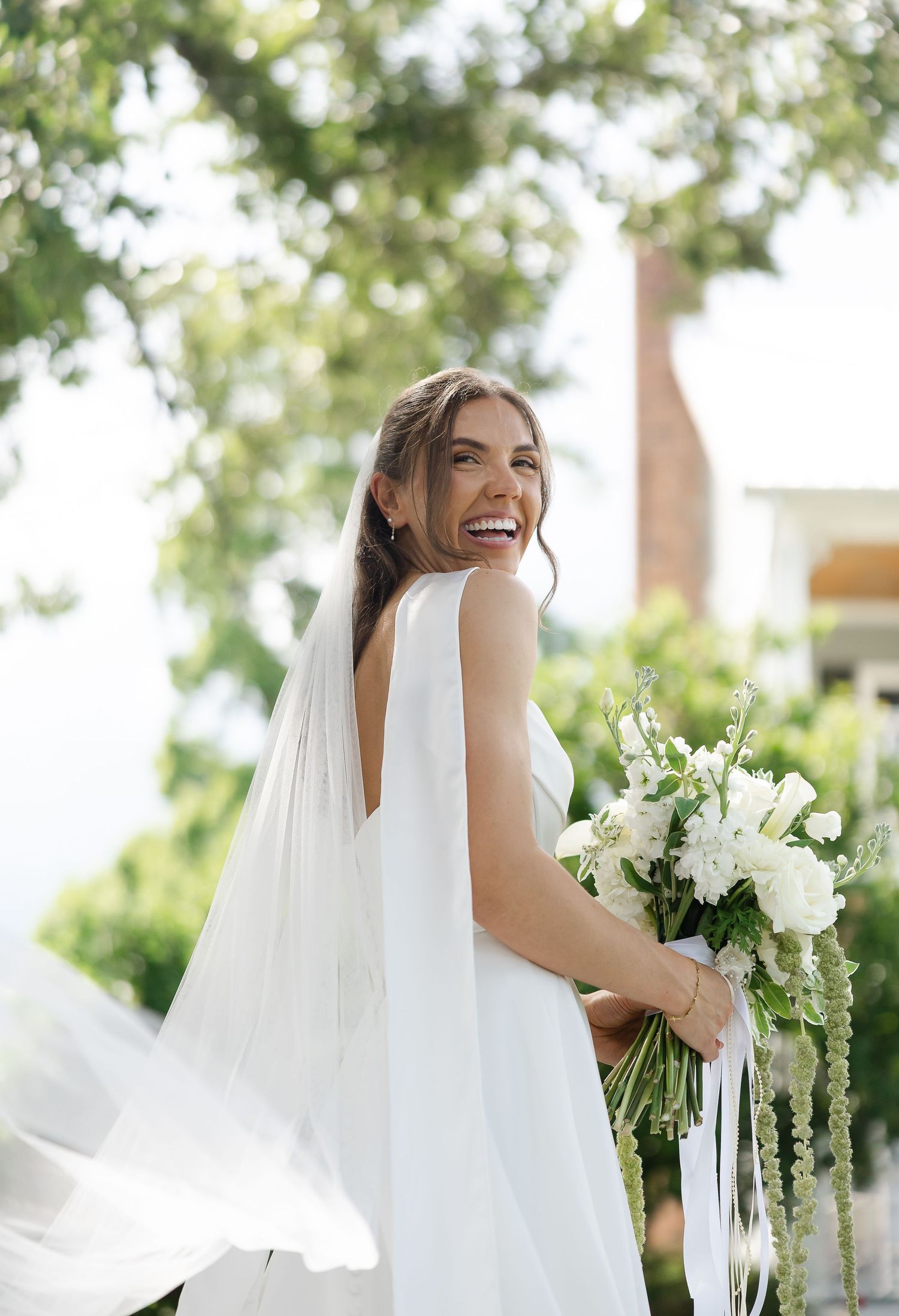 Smiling bride in white gown holding white floral bouquet outdoors with lush green trees in background.