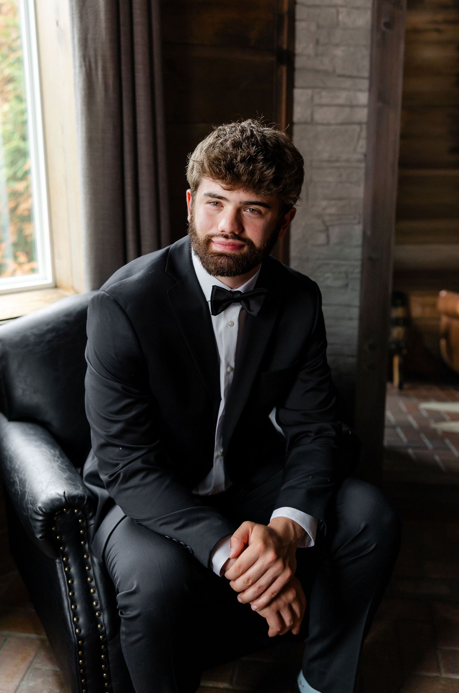 Young man with beard in black tuxedo and bow tie sitting in leather chair near window.