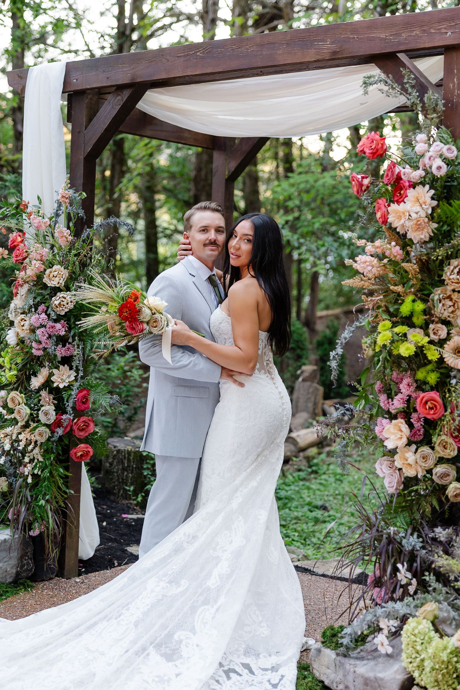 Bride and groom pose under a floral wooden arch in a forest wedding ceremony, surrounded by colorful blooms.At Drakewood Farms in Nashville,