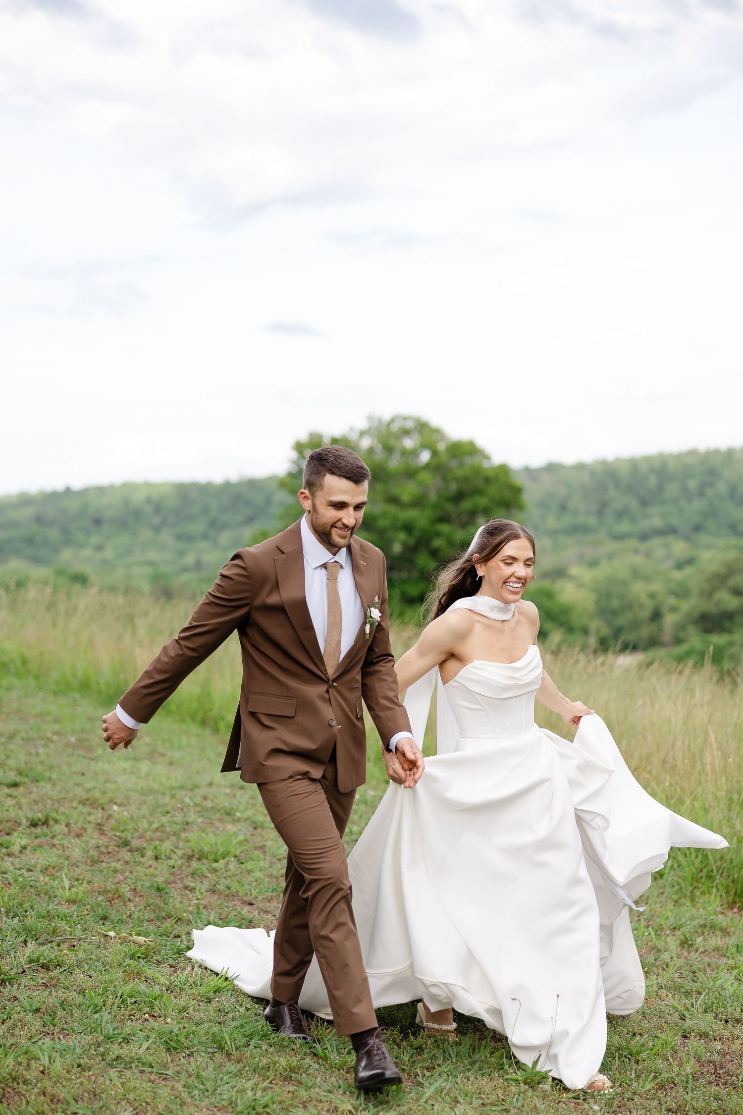 A joyful bride and groom run hand-in-hand through a lush green meadow, she lifts her white gown while he wears a brown suit.