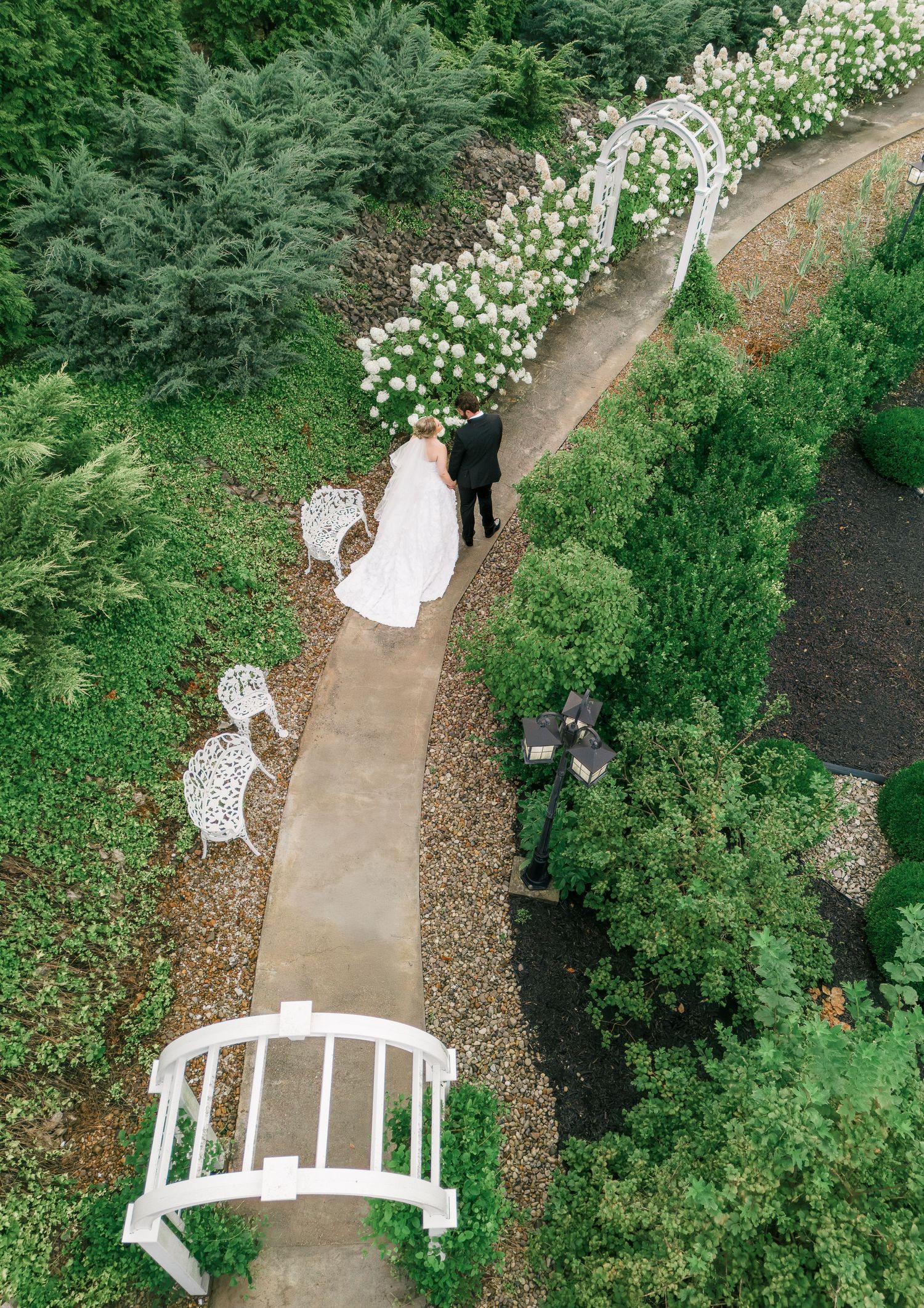 Aerial view of bride and groom walking through a lush garden pathway with white floral arbor and ornate benches.