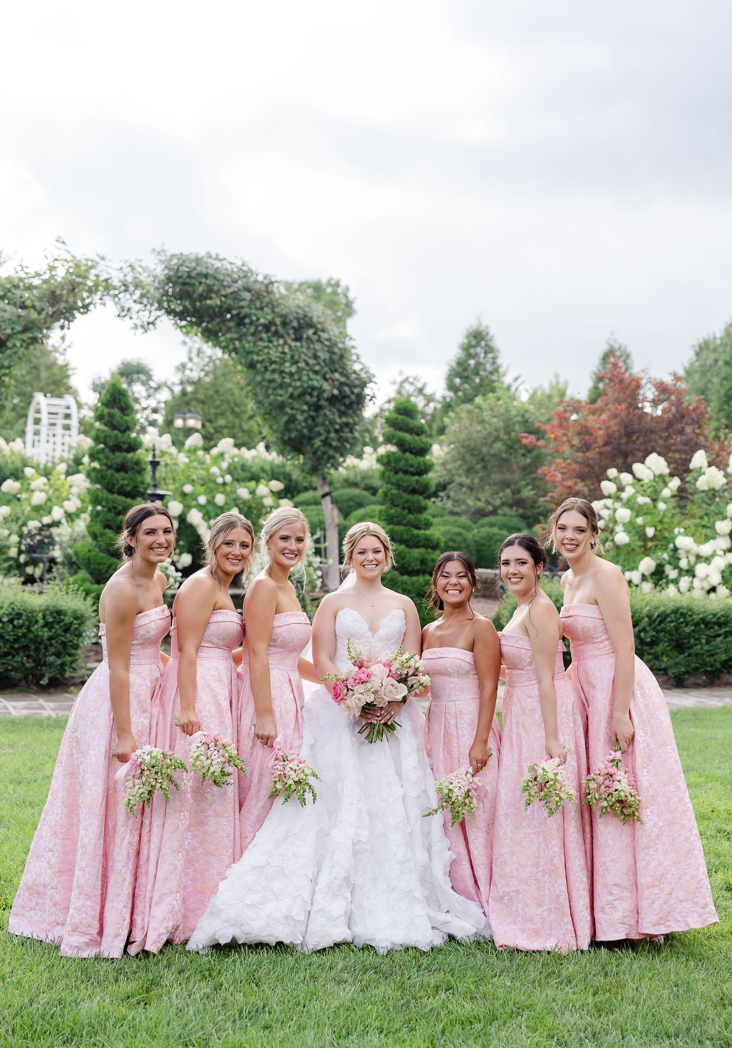 Bride in white gown poses with six bridesmaids in pink lace dresses holding bouquets in a lush garden setting.
