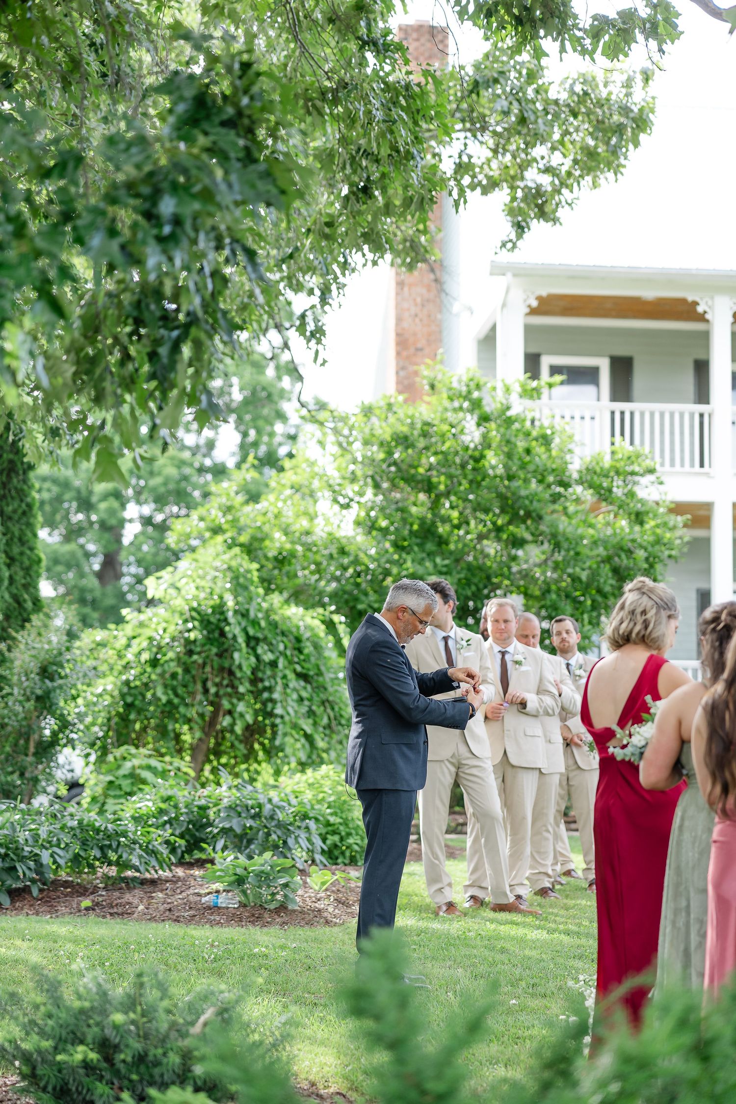 Outdoor wedding ceremony on green lawn with groomsmen in tan suits and a bridesmaid in red near a white estate.