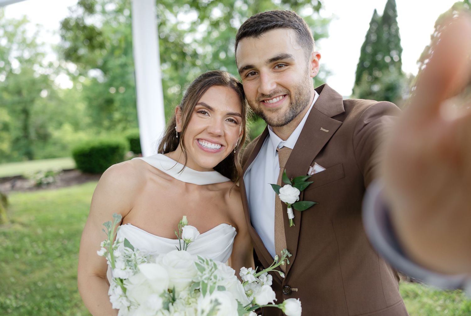 Happy newlyweds taking a selfie outdoors, bride holding white flowers in strapless gown, groom in brown suit with boutonniere.