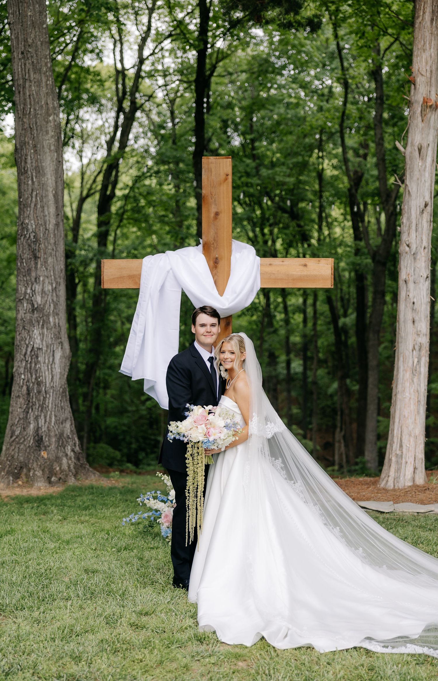 Bride and groom pose before a wooden cross draped in white fabric, surrounded by lush green trees.
