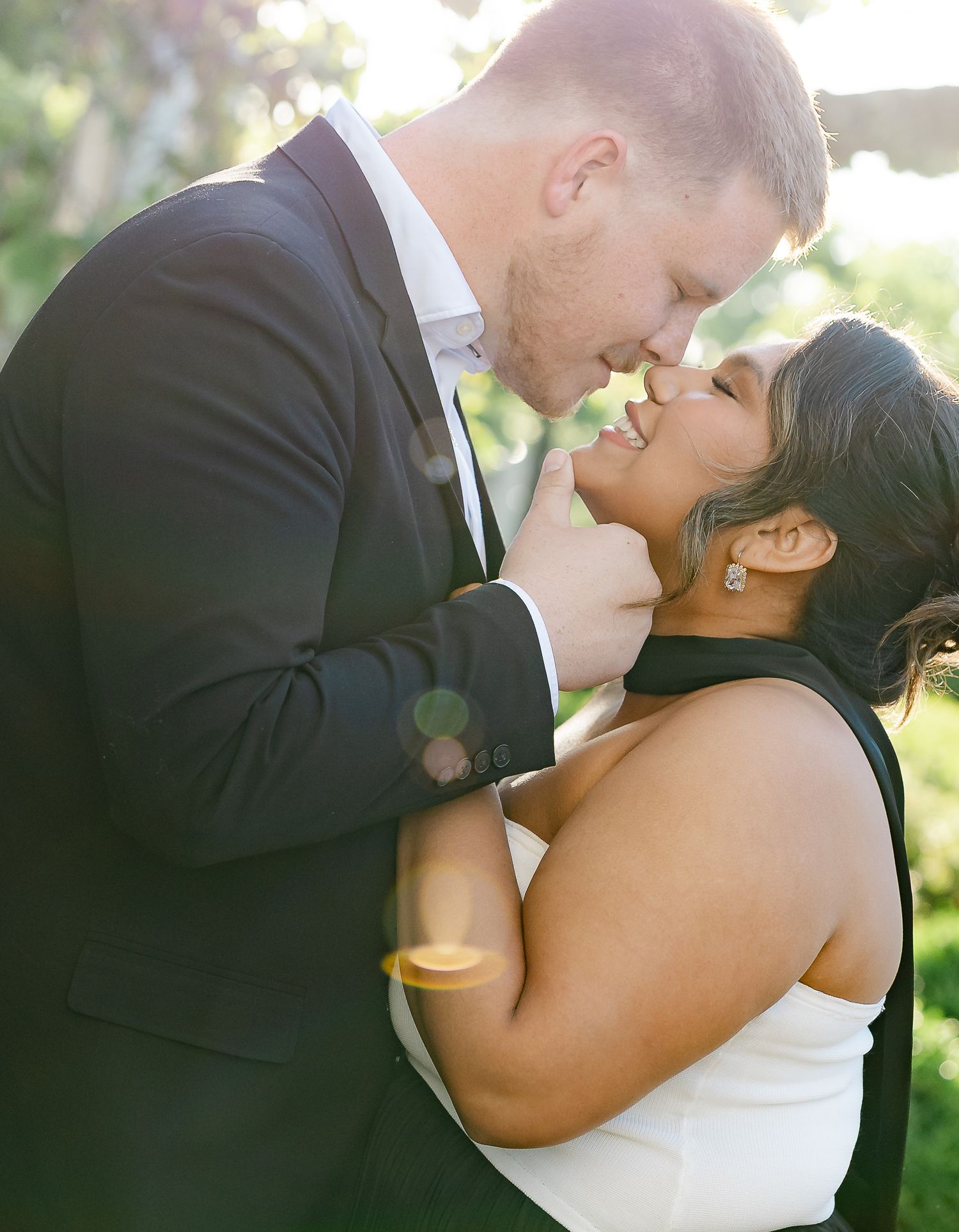 A couple shares a romantic moment outdoors, the groom in a dark suit leaning in toward his smiling bride in a white dress.