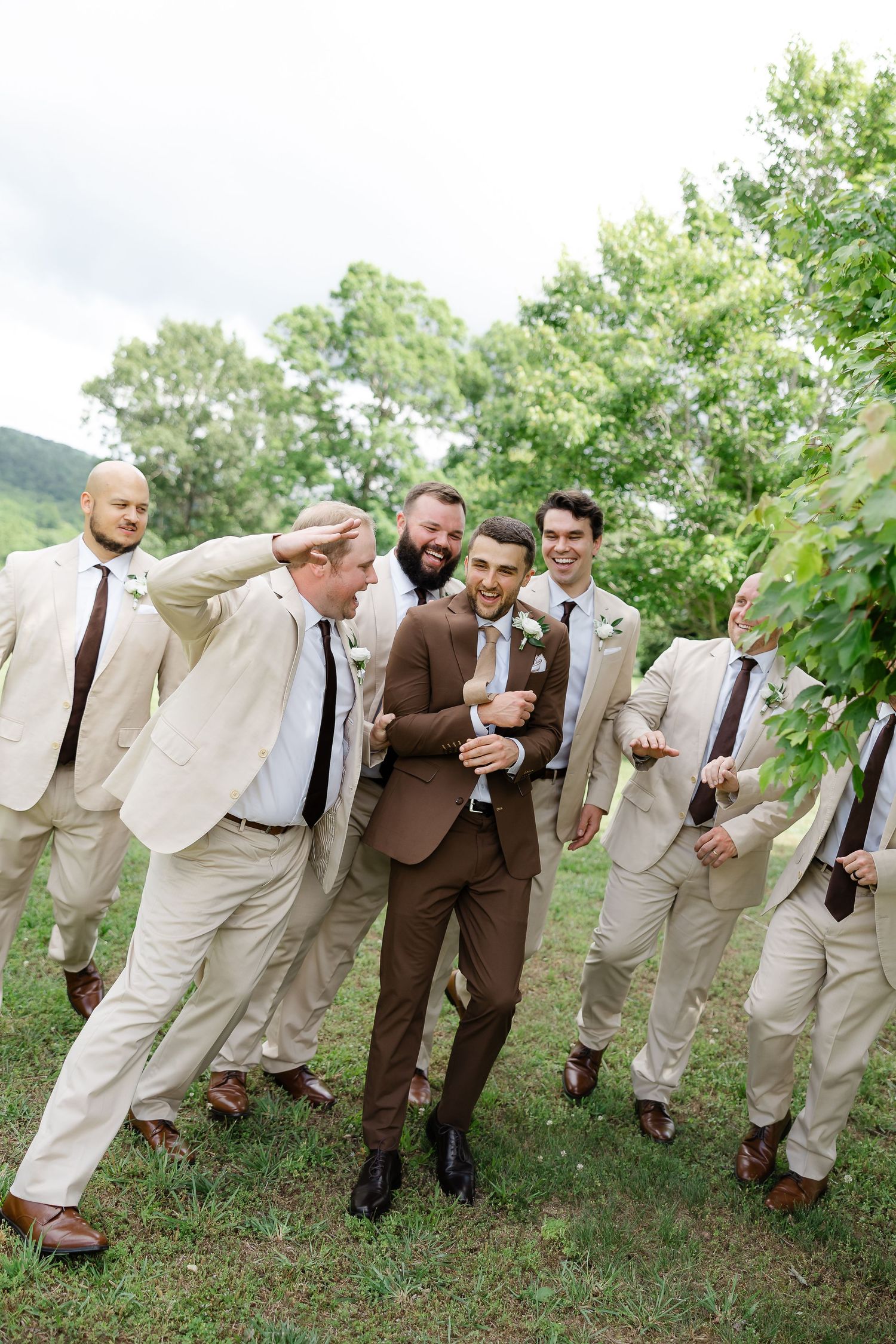 Groom in brown suit laughing with groomsmen in tan suits and dark ties outdoors at a wedding.
