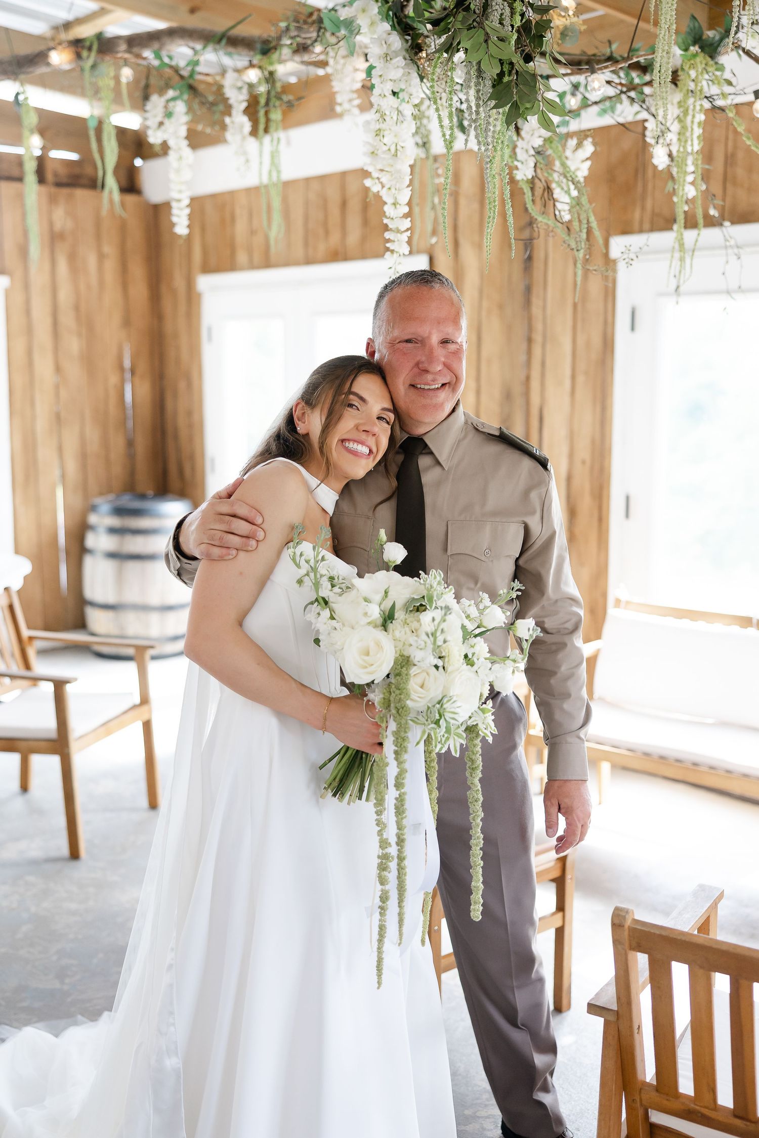 Bride in white gown holding white floral bouquet poses with older man in tan uniform inside rustic wood wedding venue.