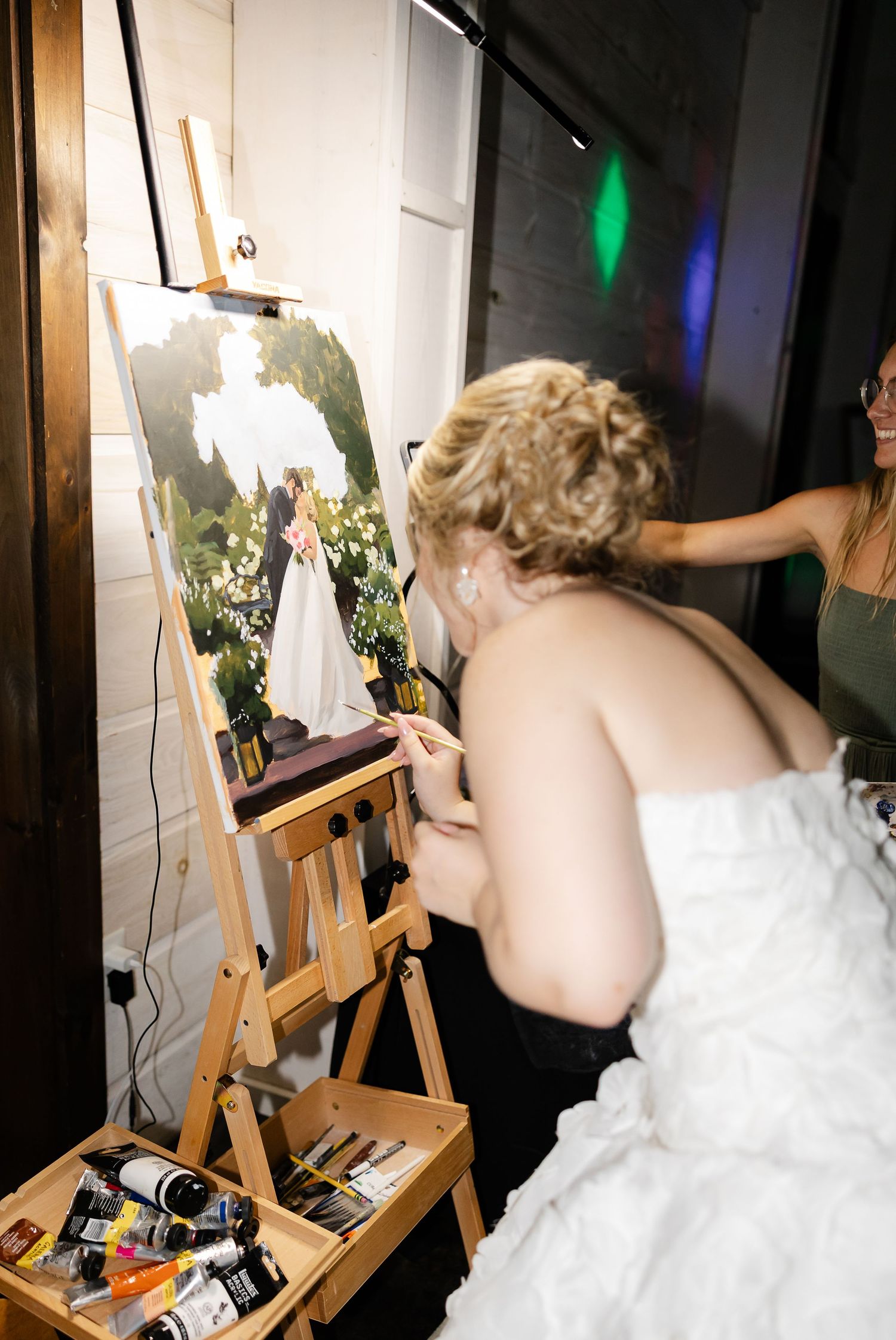 Bride in white gown painting a wedding portrait on an easel at her reception.