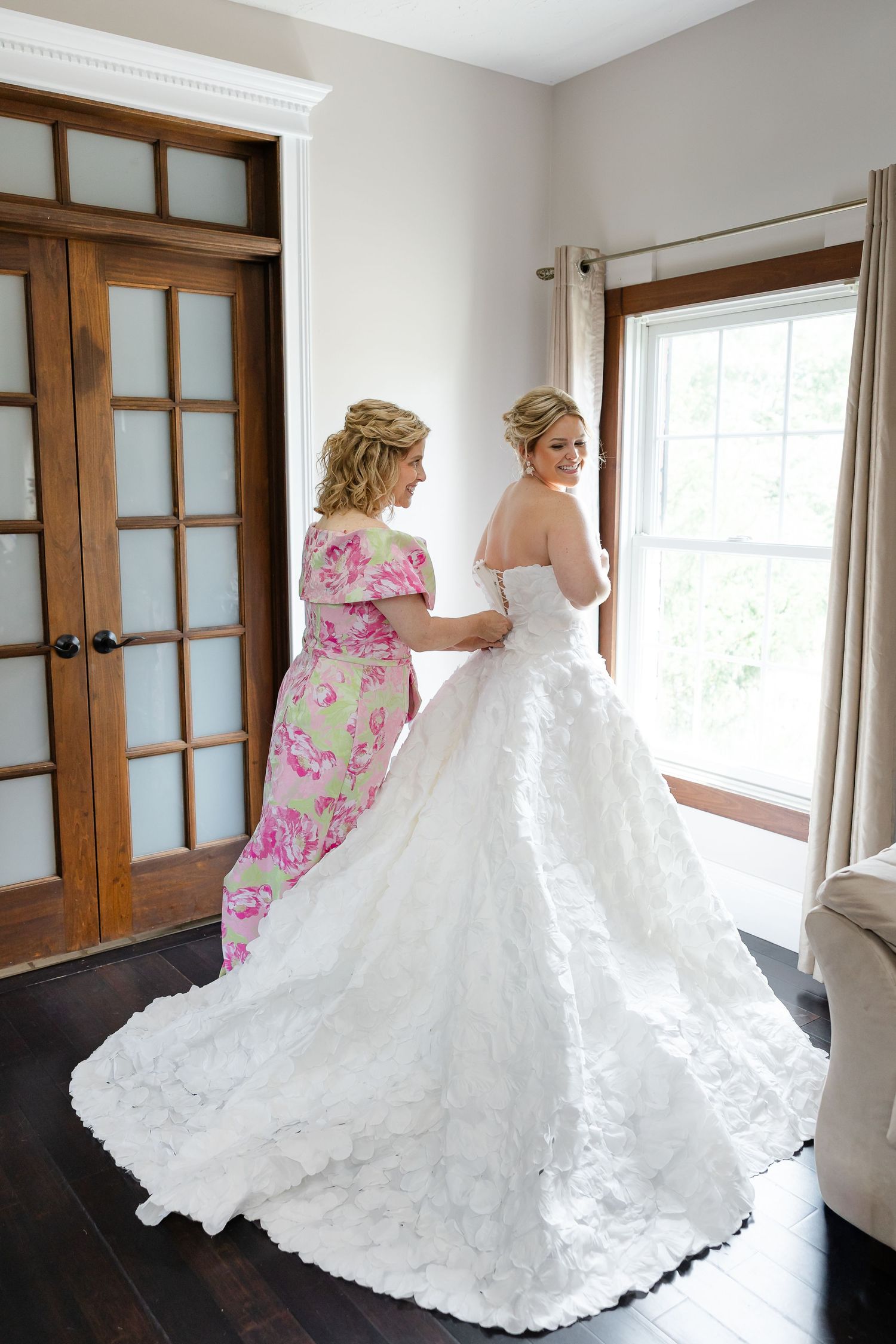 Bride in white lace ball gown being helped by woman in floral dress near window in elegant bridal suite.