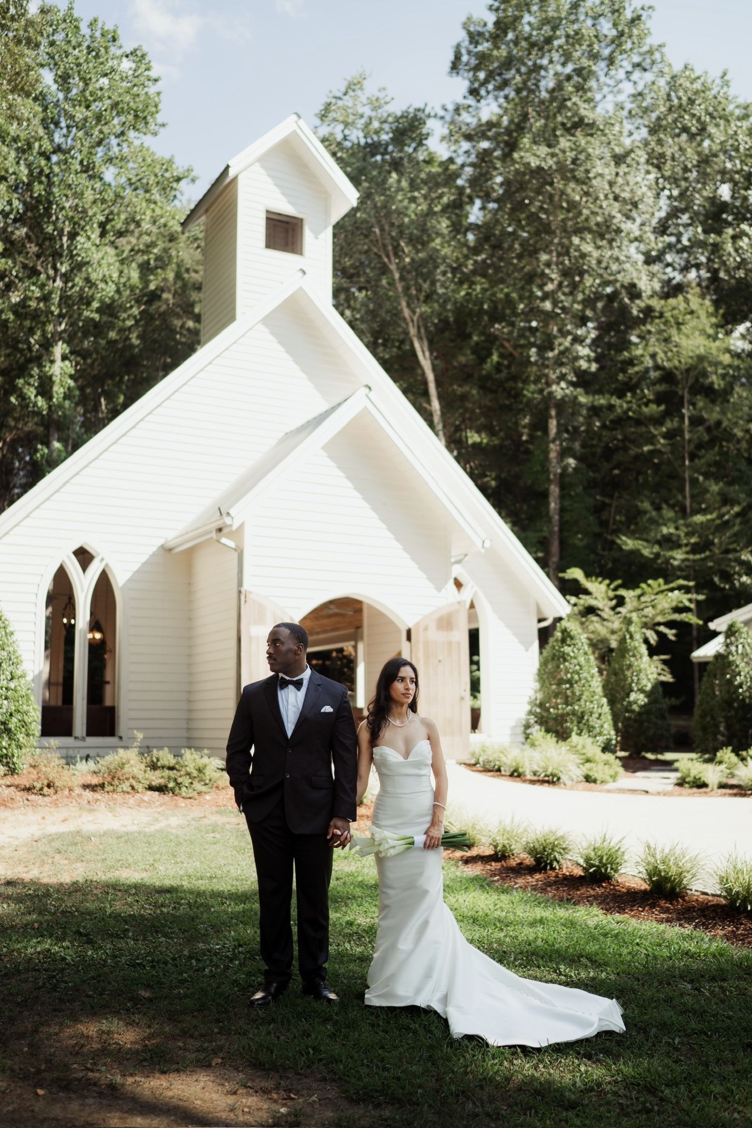 Bride and groom holding hands in front of a white chapel surrounded by lush green trees at FireFly Lane Chapel located in Dickson Tennessee