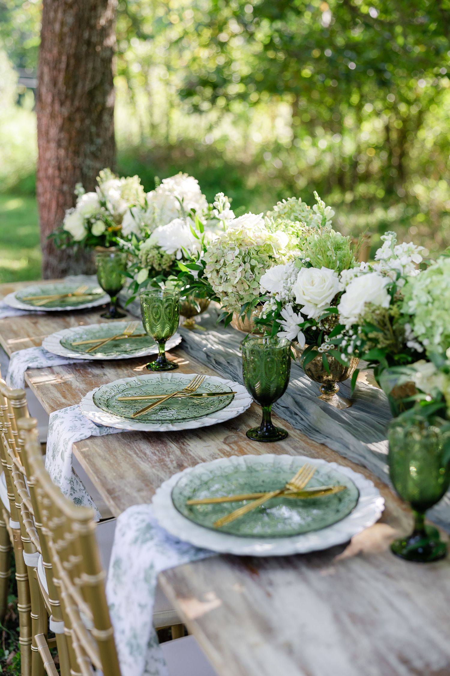 Elegant outdoor wedding tablescape with green glass plates, gold cutlery, white floral centerpieces, and emerald goblets on rustic wood.
