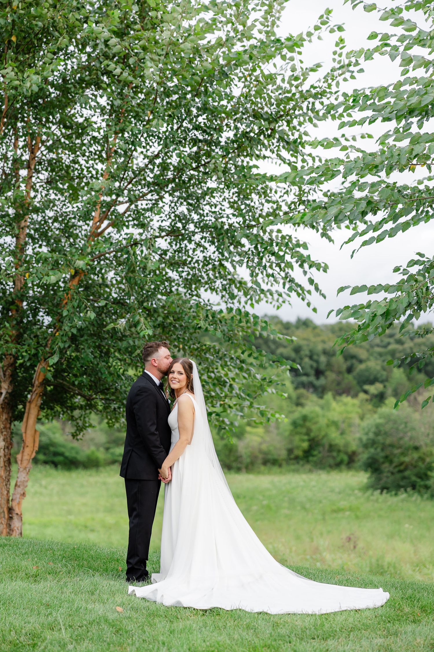 Bride and groom share a romantic moment under a lush green tree on their wedding day in a scenic outdoor setting.At The Venue at Birchwood