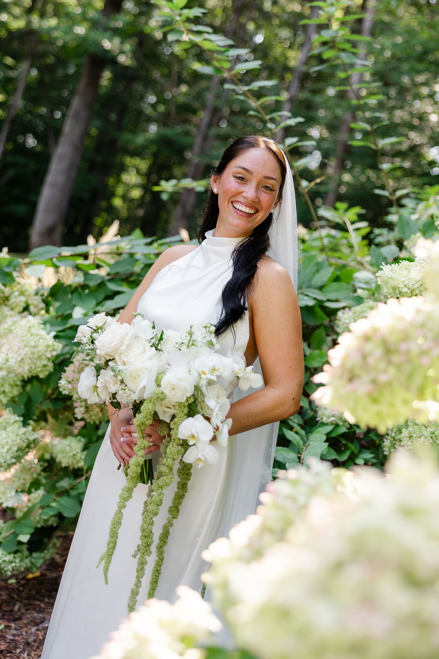 Smiling bride in white halter gown holds white floral bouquet surrounded by lush green hydrangeas outdoors At FireFly Lane in Dickson, TN
