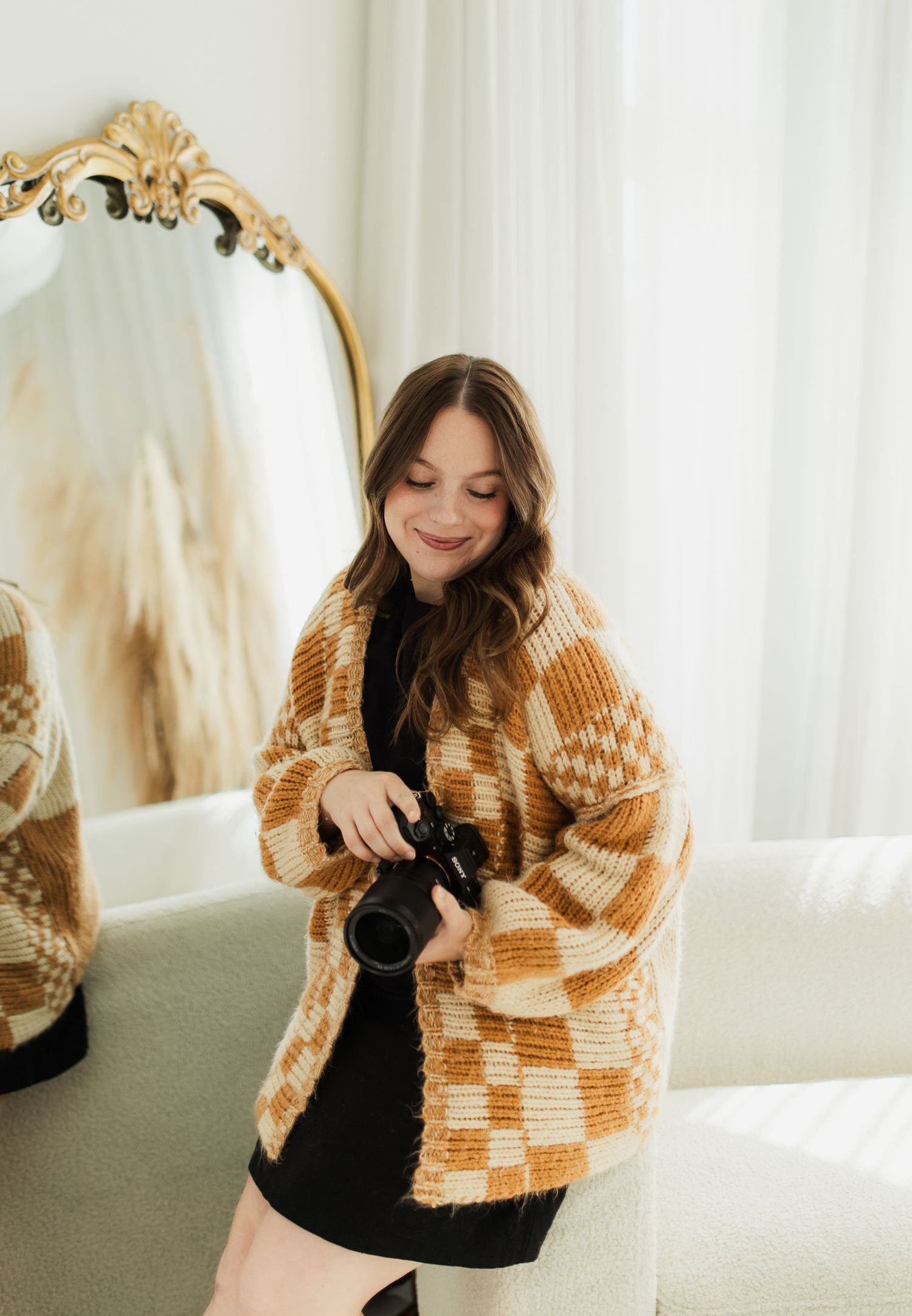 Young woman in checkered cardigan holding a camera, smiling in a bright room with a gold ornate mirror.