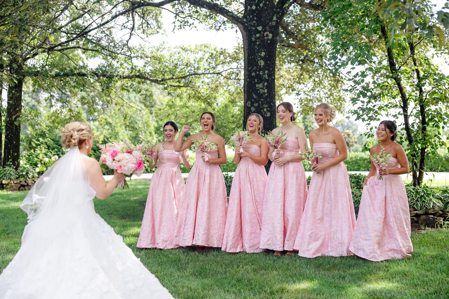Bride in white gown faces six bridesmaids in pink dresses holding bouquets outdoors under a large tree.