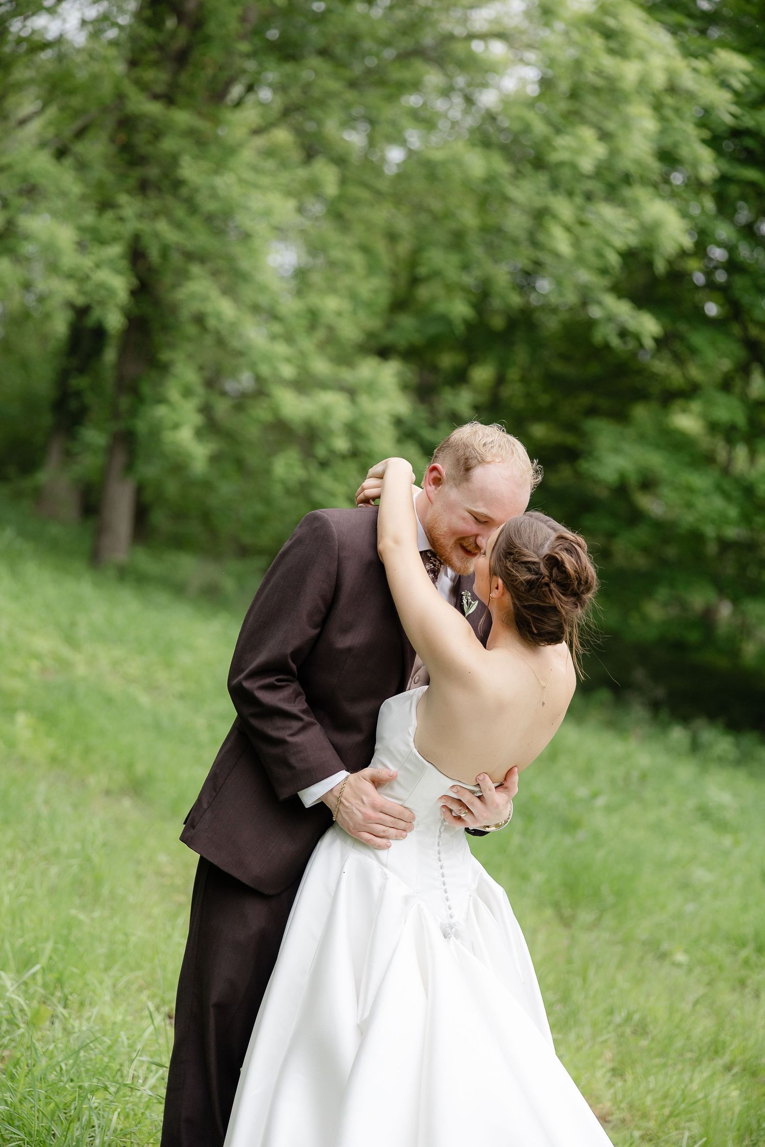 A bride and groom share a romantic kiss outdoors, surrounded by lush green trees, as she wears a backless white gown.