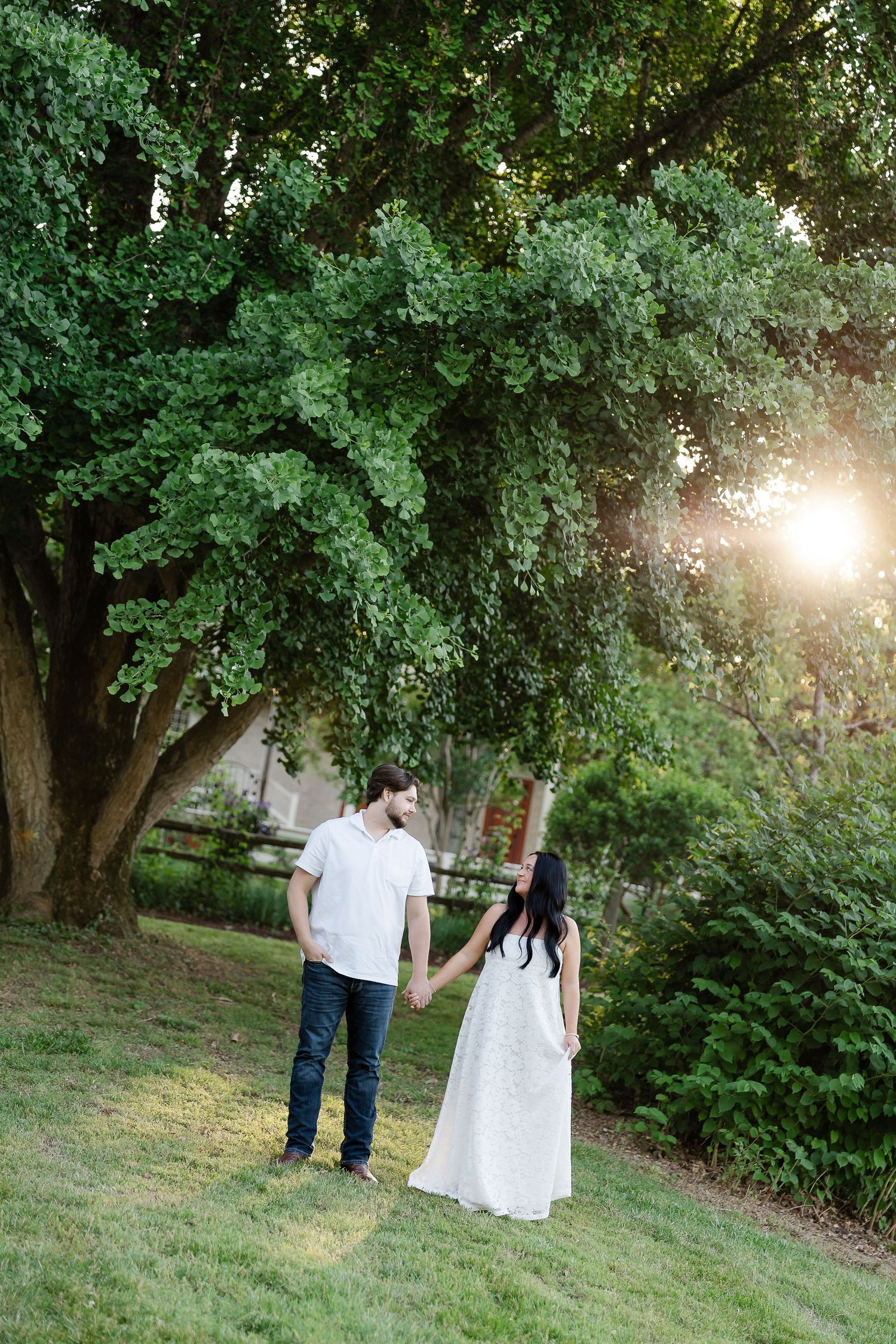 Couple holding hands in white outfits under a large green tree with golden sunlight in a lush garden.