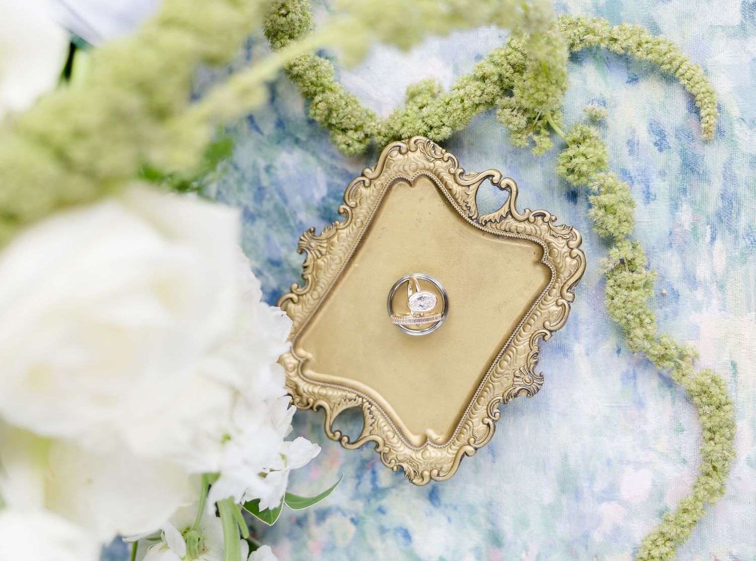 Gold ornate ring dish holding a diamond engagement ring surrounded by white flowers and green moss on a blue background.