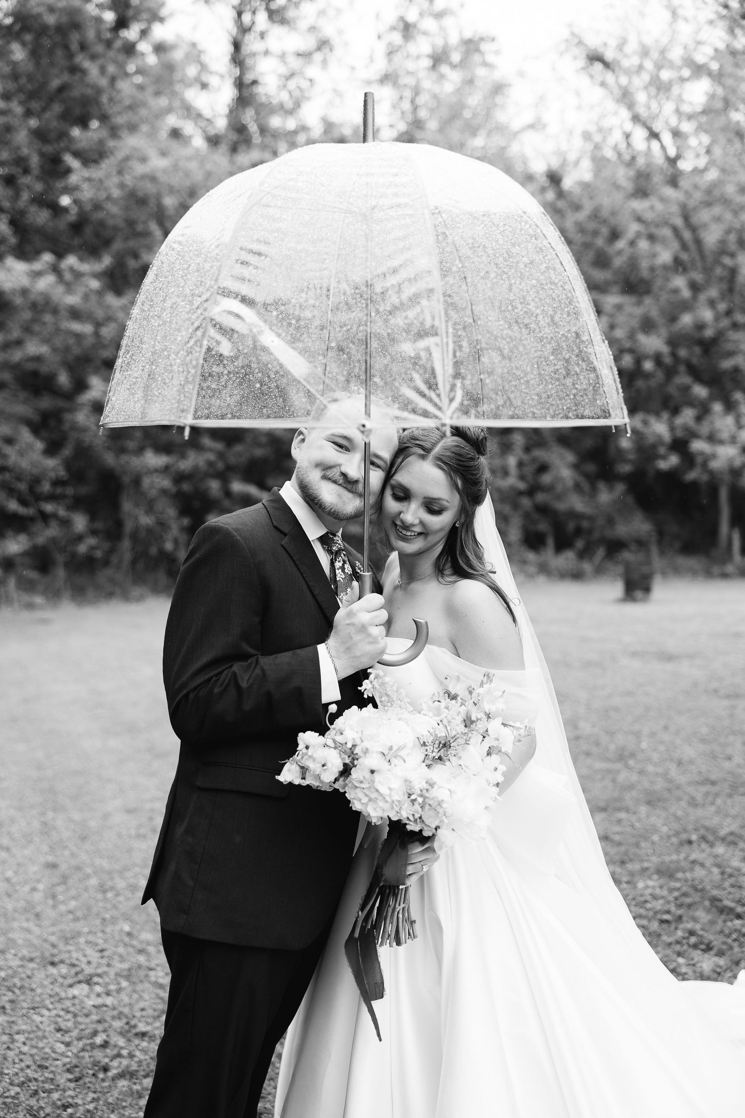 Black and white photo of a bride and groom smiling under a clear dome umbrella, bride holding a floral bouquet.