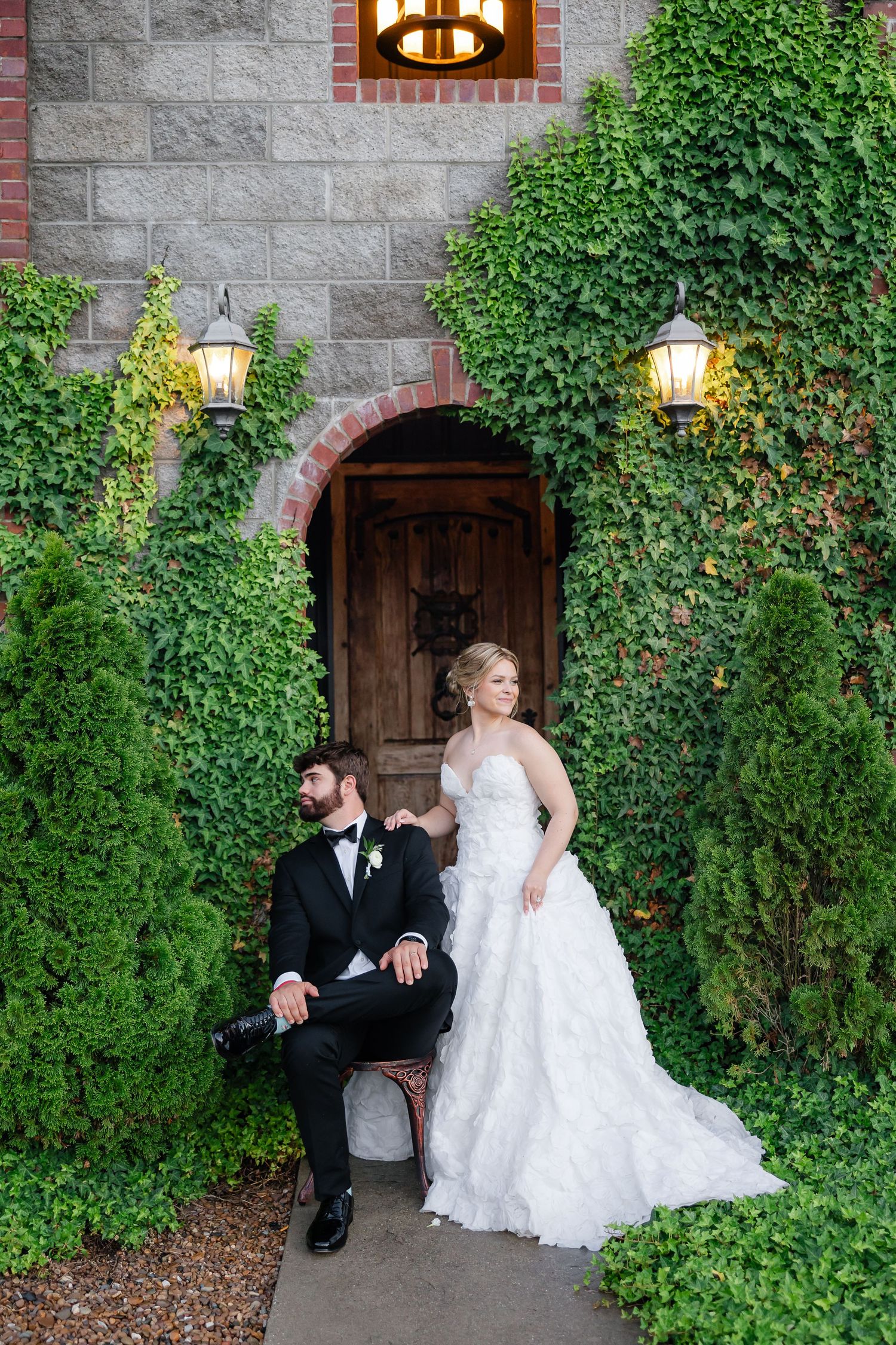 Bride in white ruffled gown stands beside seated groom in tuxedo before ivy-covered stone building with arched wooden door.