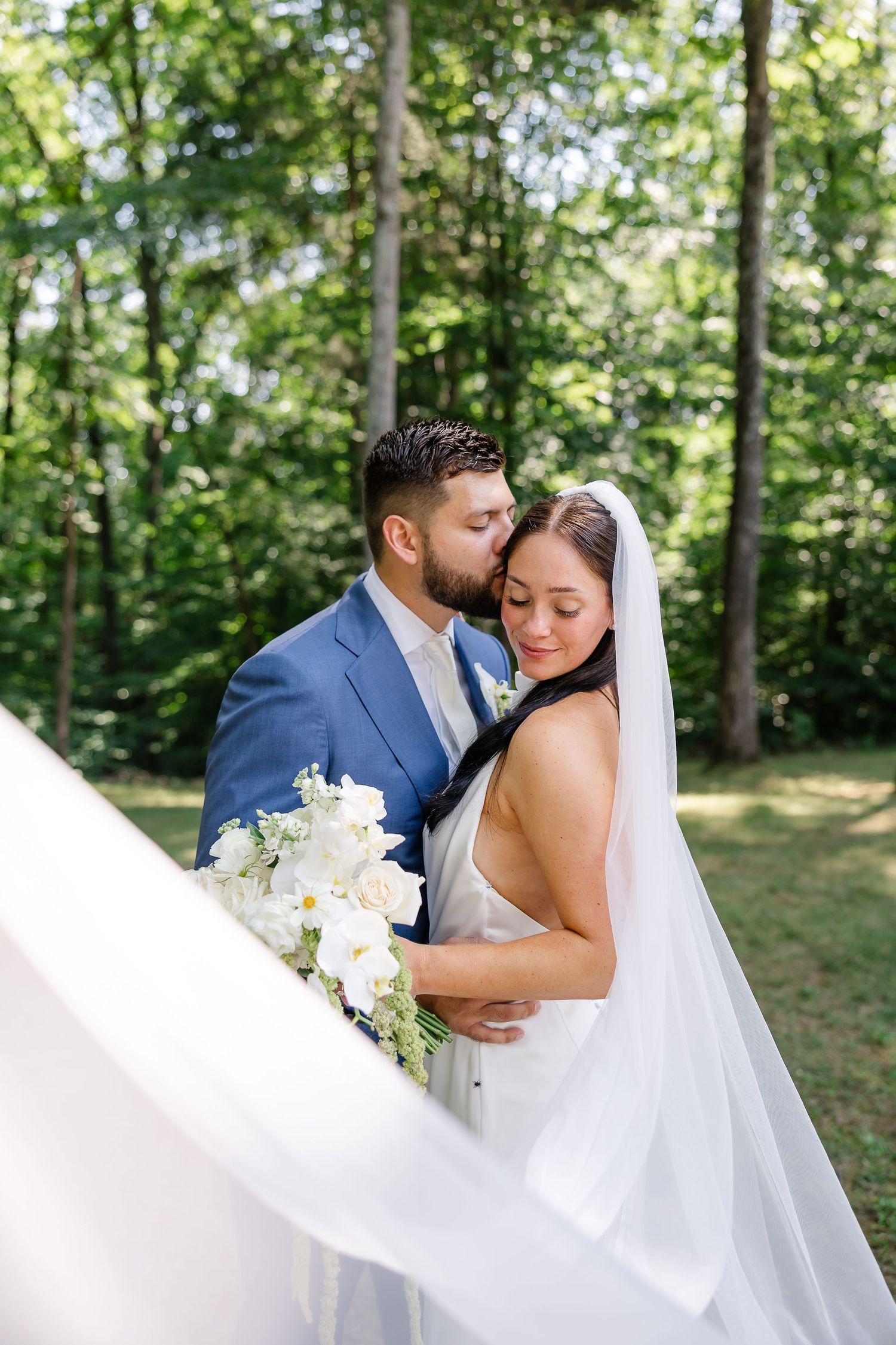 Groom in blue suit kisses bride wearing white veil and gown while she holds white floral bouquet outdoors. At FireFly Lane in Dickson, TN