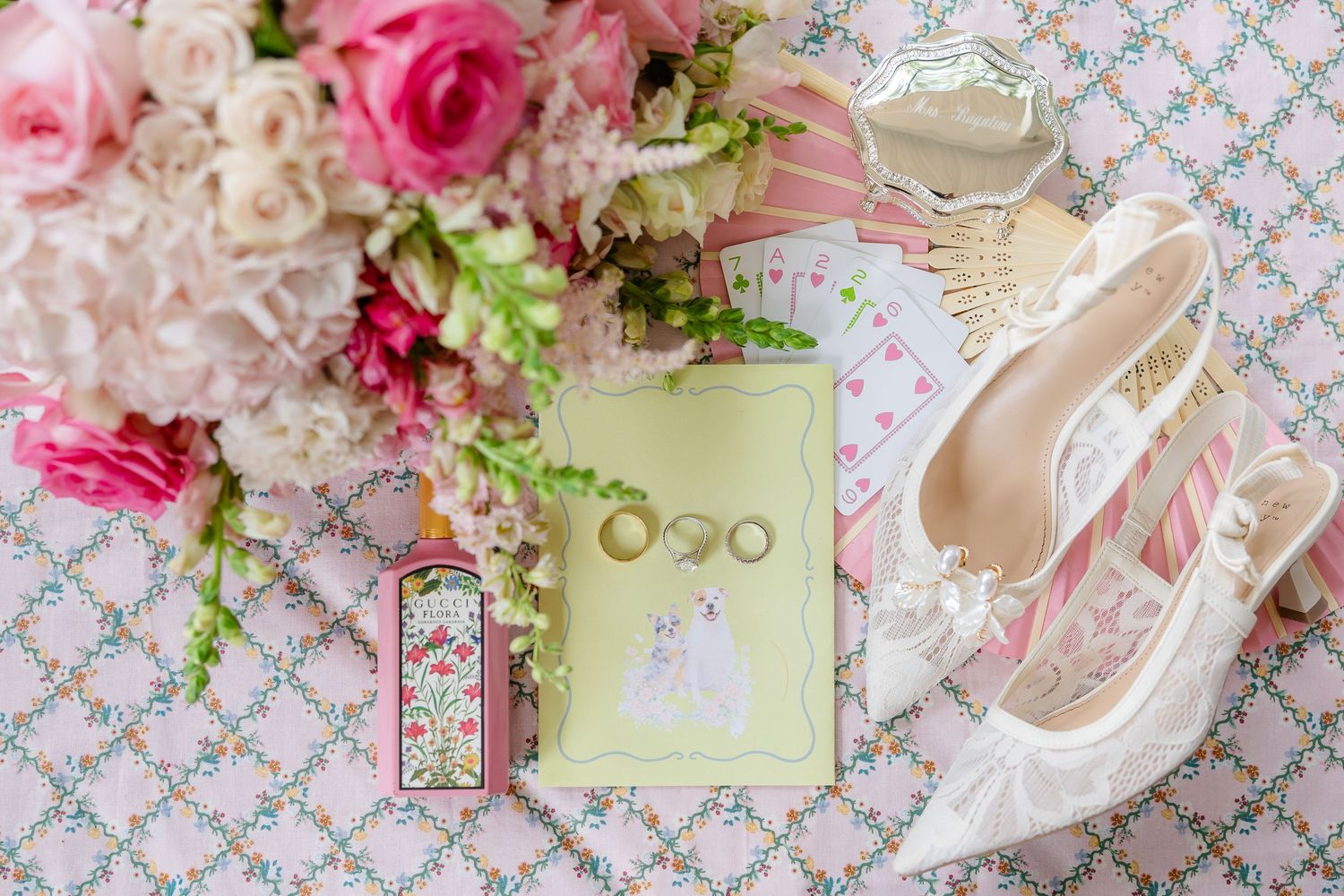 Elegant bridal flat lay featuring pink roses, wedding rings, lace heels, pearl jewelry, and a green invitation card on floral fabric.