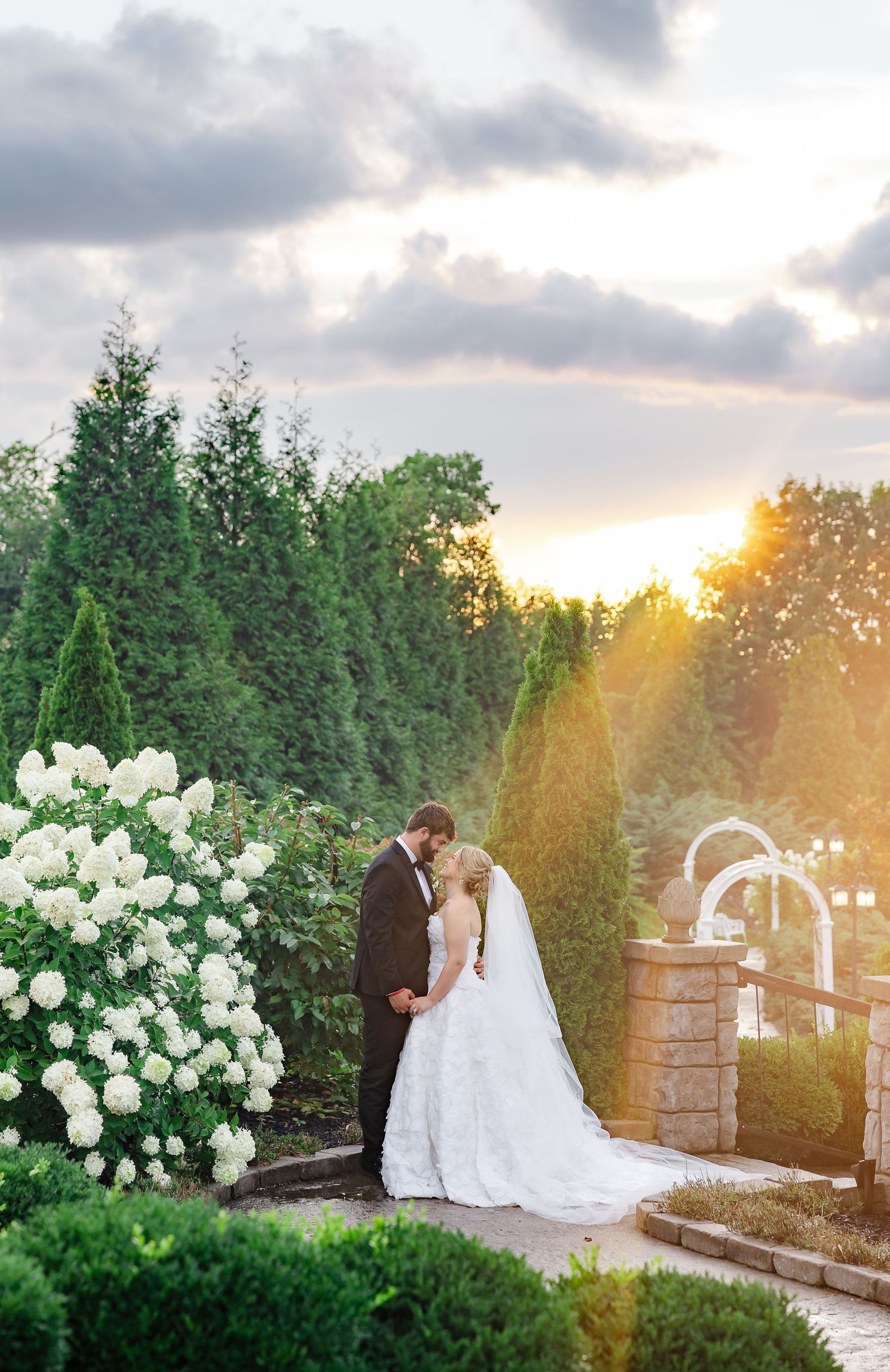 Bride and groom share a romantic sunset kiss surrounded by lush greenery and white hydrangeas in a garden.