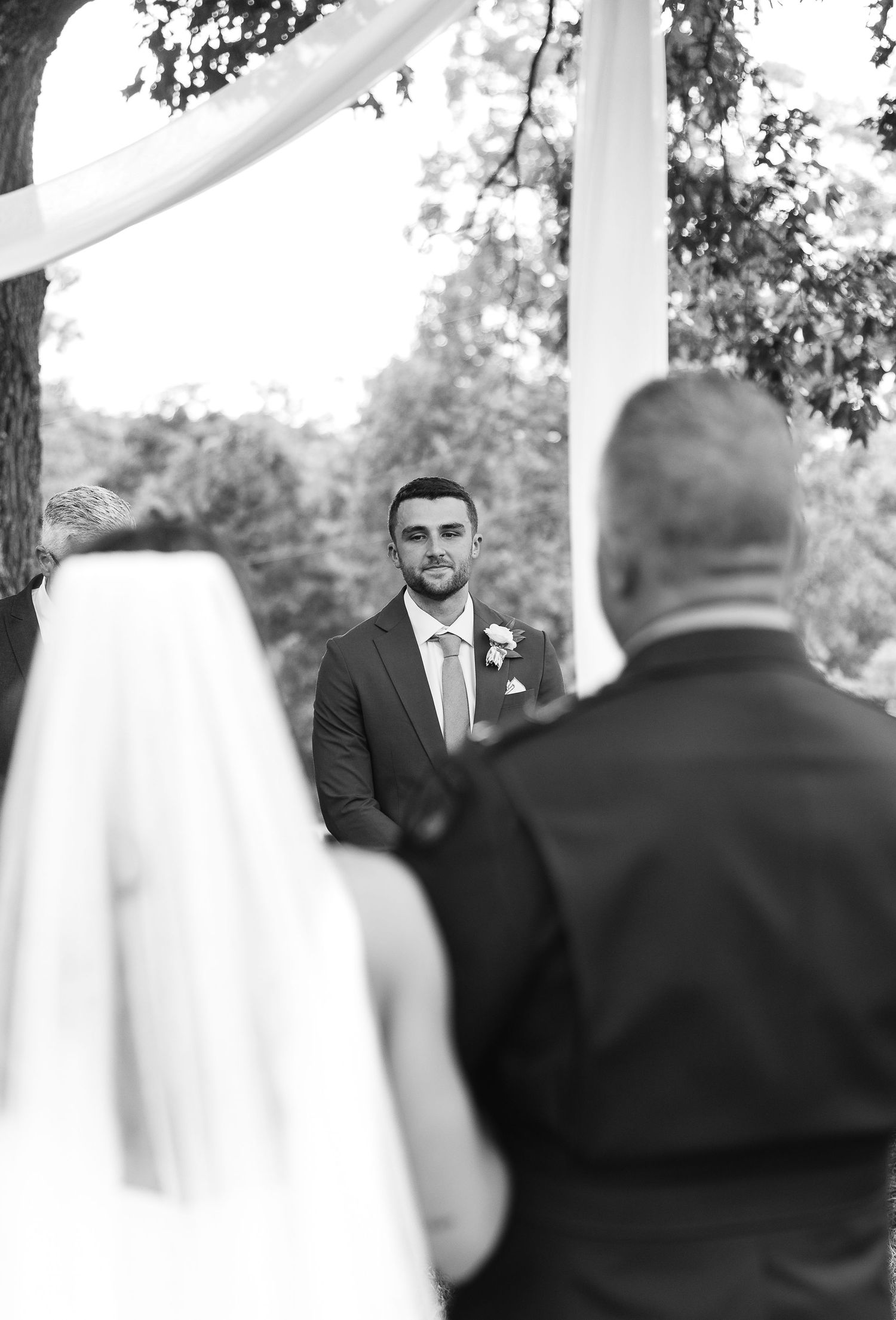 Groom watches bride walk down the aisle at outdoor wedding ceremony in black and white photo.